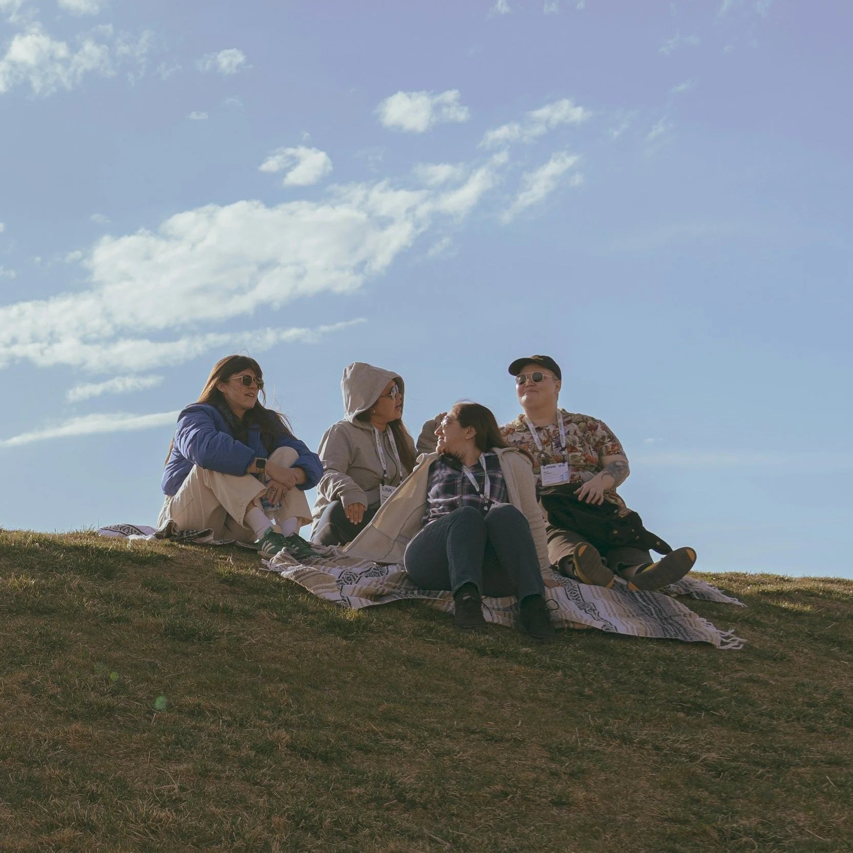 A group of five people sitting on a blanket on a hillside, outdoors, with a bright blue sky and clouds above. They are wearing casual clothing and sunglasses, and appear to be enjoying a sunny day together.