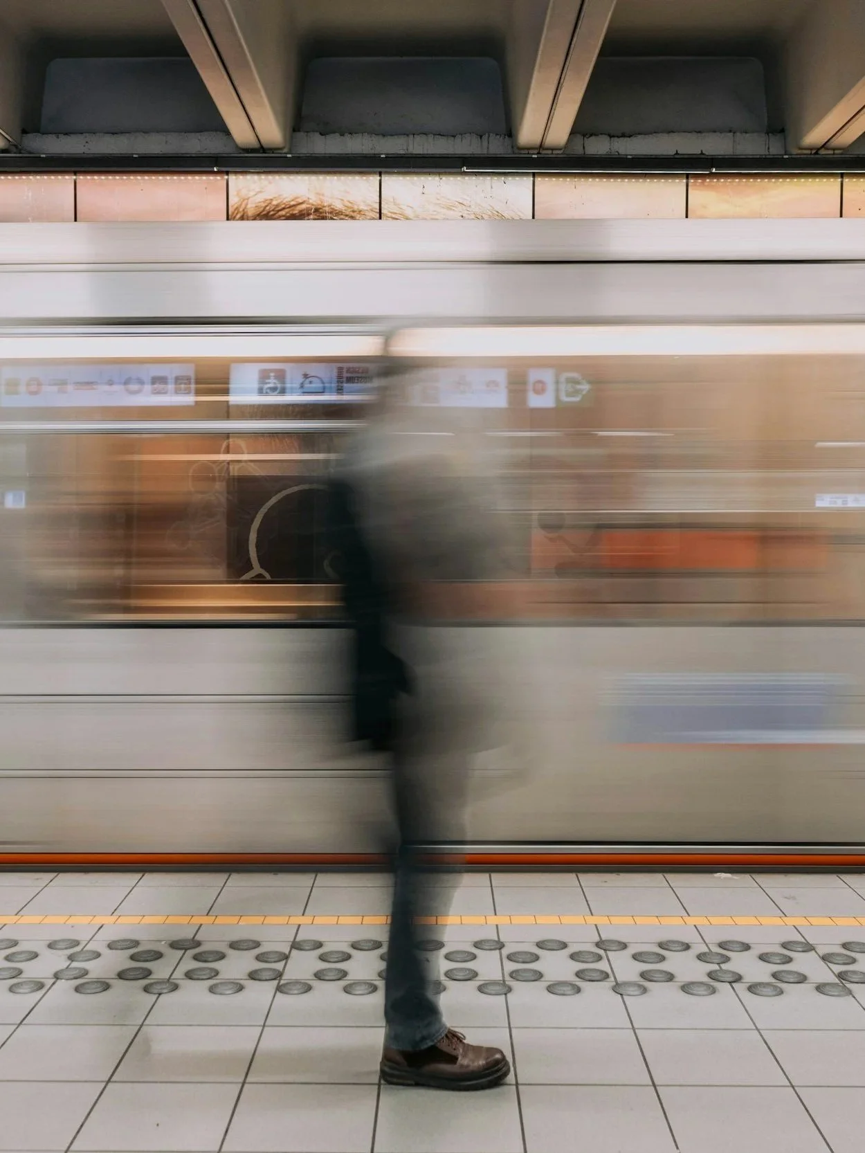 A blurry image of a person standing on a subway platform with a moving train passing in front of them. The person is wearing dark clothing and brown shoes.