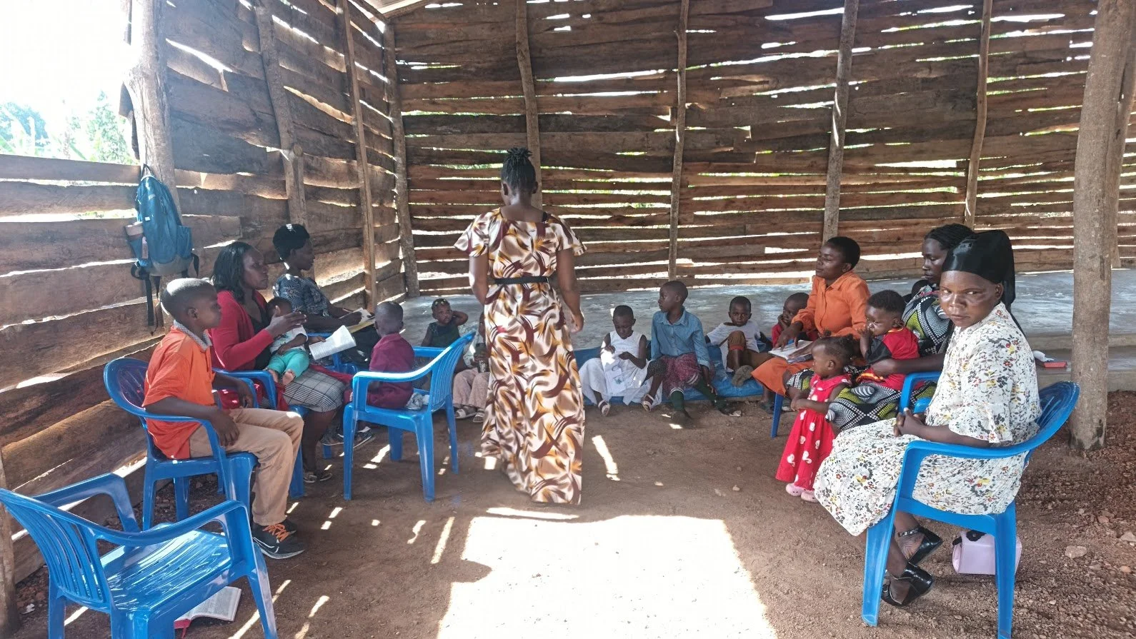 Inside a rustic wooden structure with children and adults seated on blue plastic chairs and a woman standing in front. The walls are made of horizontal wooden planks with gaps, and the ground is dirt. Sunlight filters through the gaps in the walls.