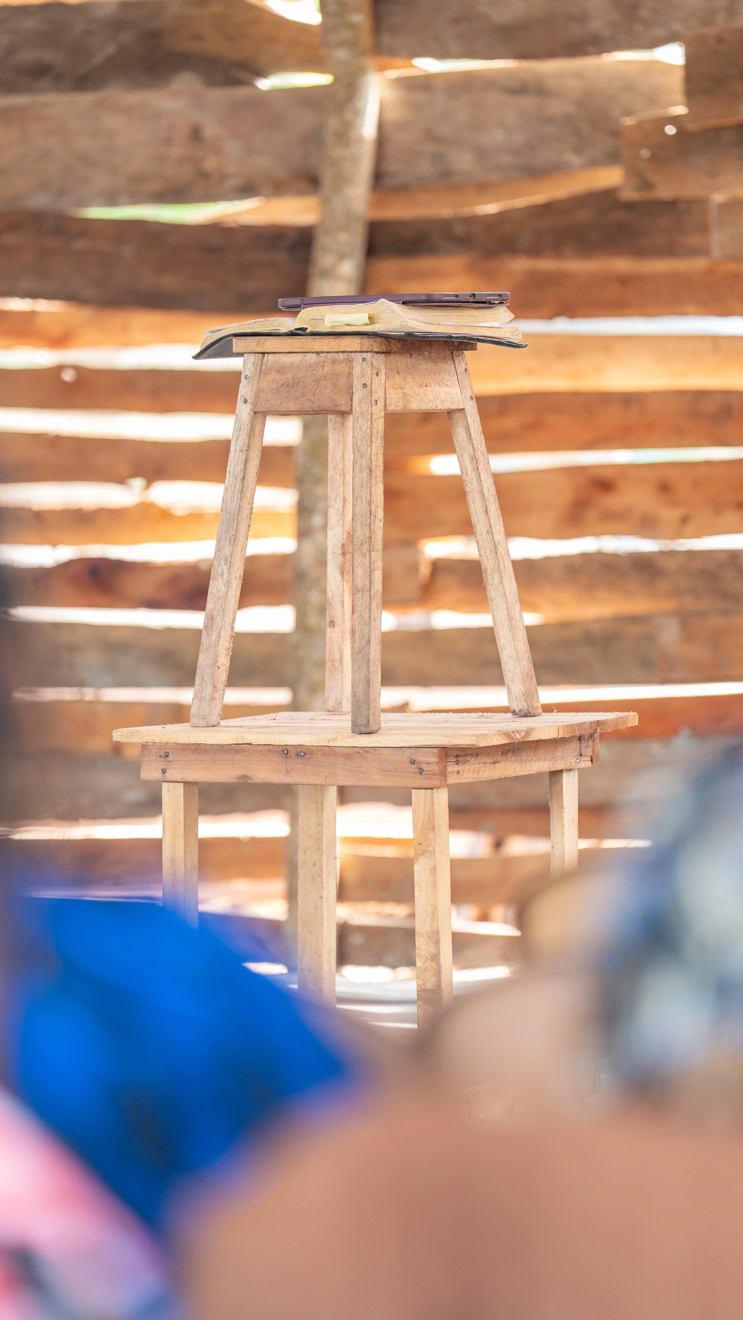A wooden chair with a small wooden table on top, placed inside a rustic room with a wooden plank wall.