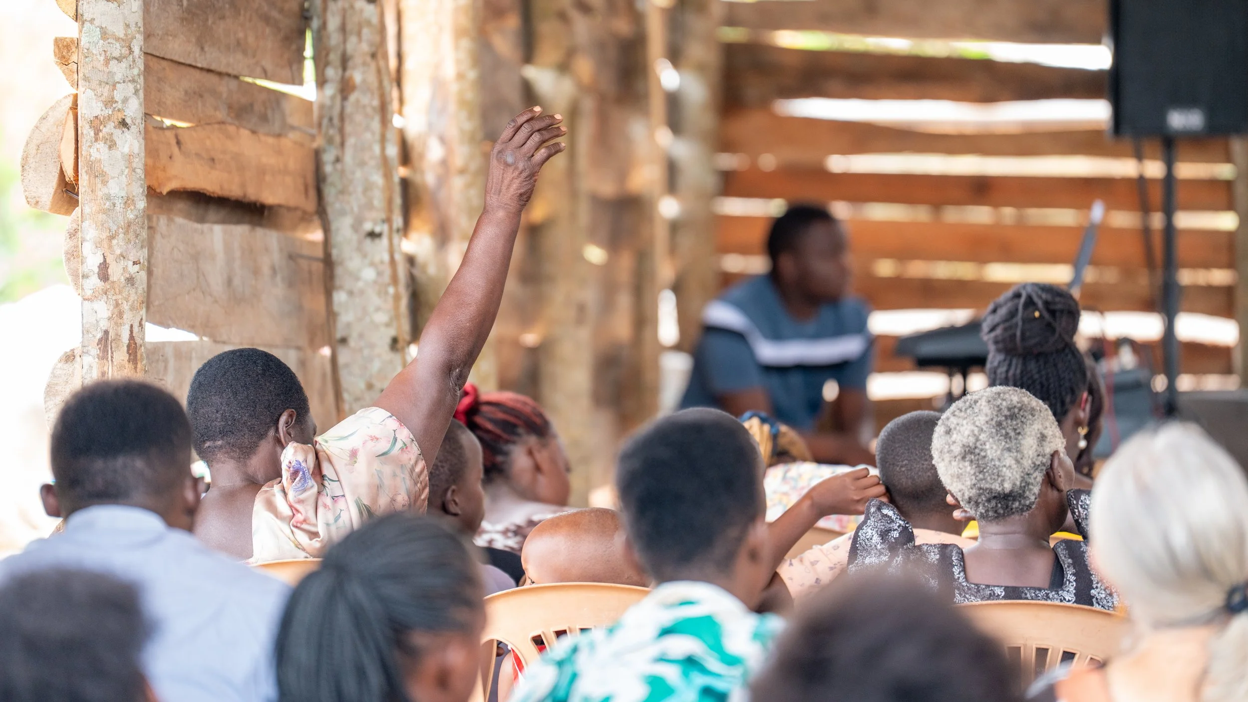 A group of children and adults sitting in a rustic wooden room, with one child raising their hand during a class or presentation, with a person at the front near a computer.