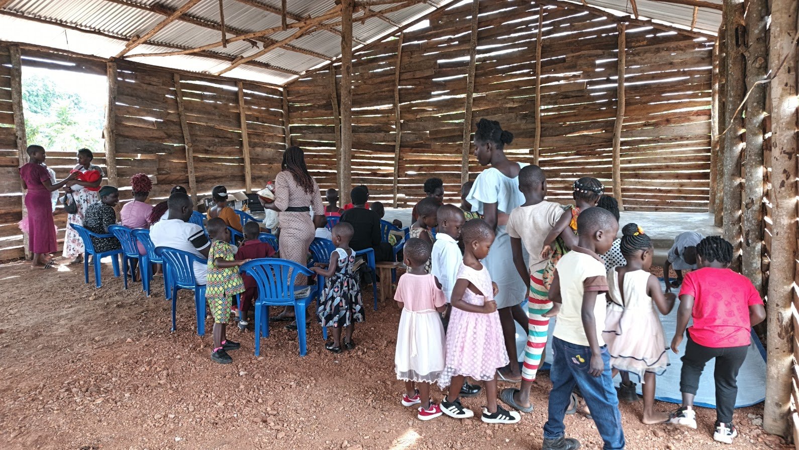 Children and adults inside a rustic wooden building, some seated on blue chairs and others standing, engaged in activities or conversations during a community event.
