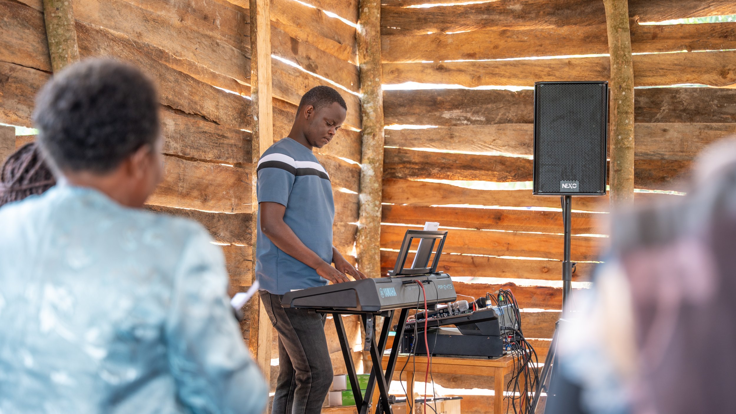 A young man playing a keyboard at an indoor event with a rustic wooden interior, with a large speaker nearby and audience members seated around.