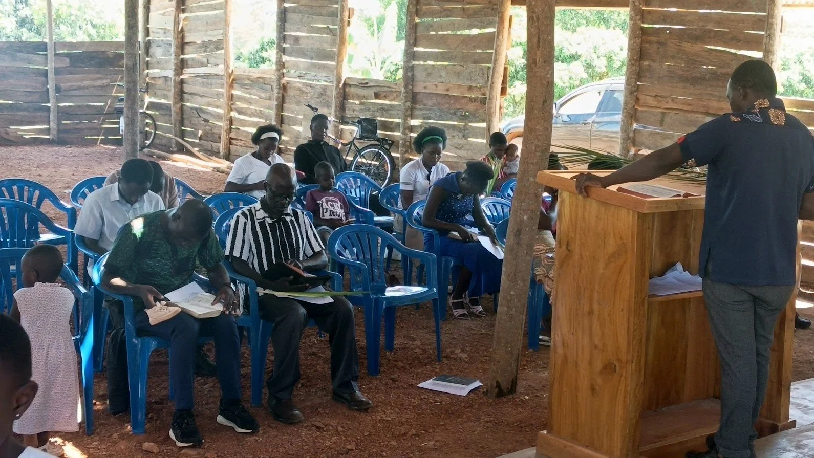 A church gathering with men, women, and children seated on blue plastic chairs inside a rustic wooden structure, with a pastor addressing the congregation from a wooden pulpit.