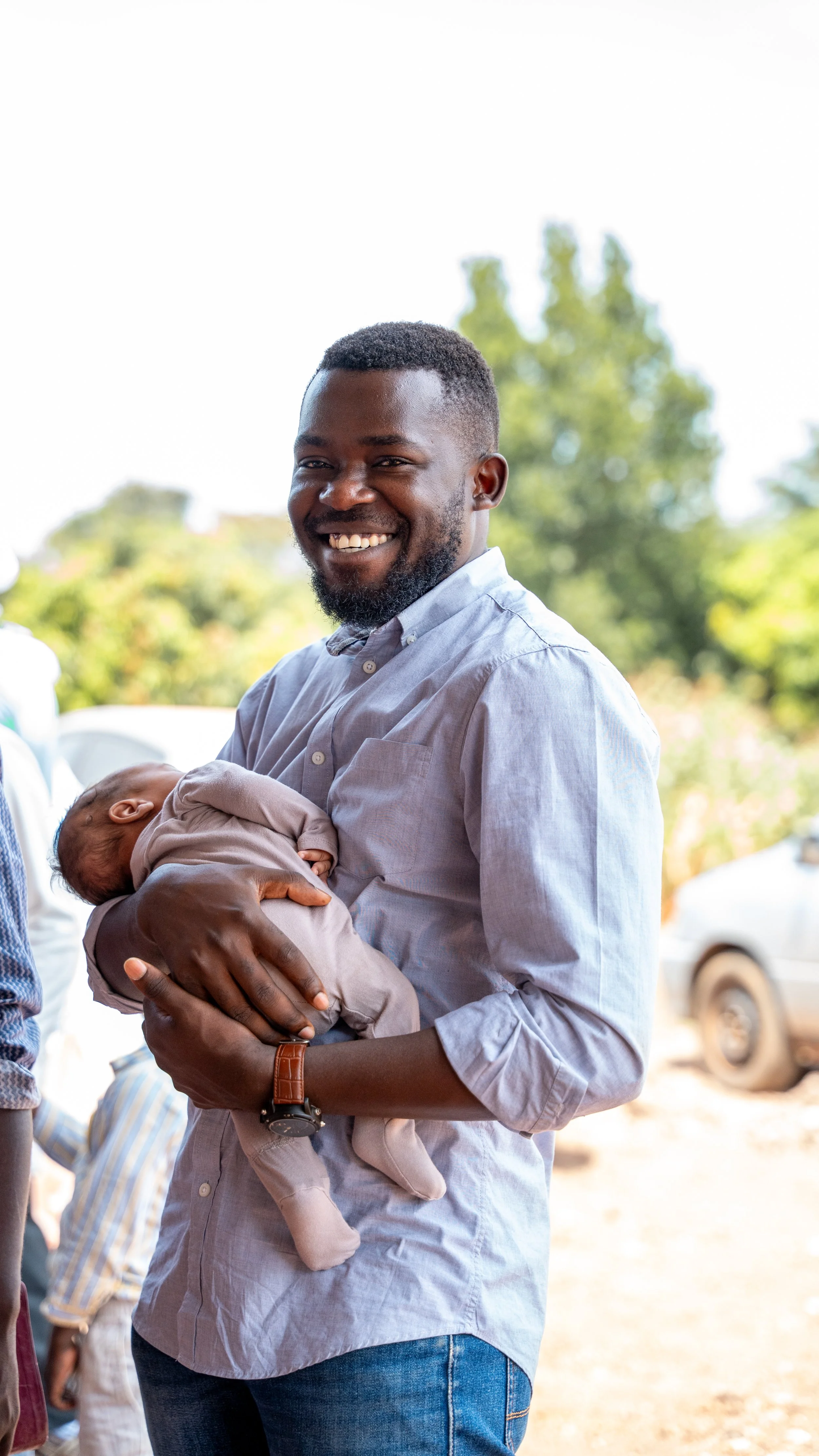 A smiling man holding a sleeping baby outside during daytime with trees and cars in the background.