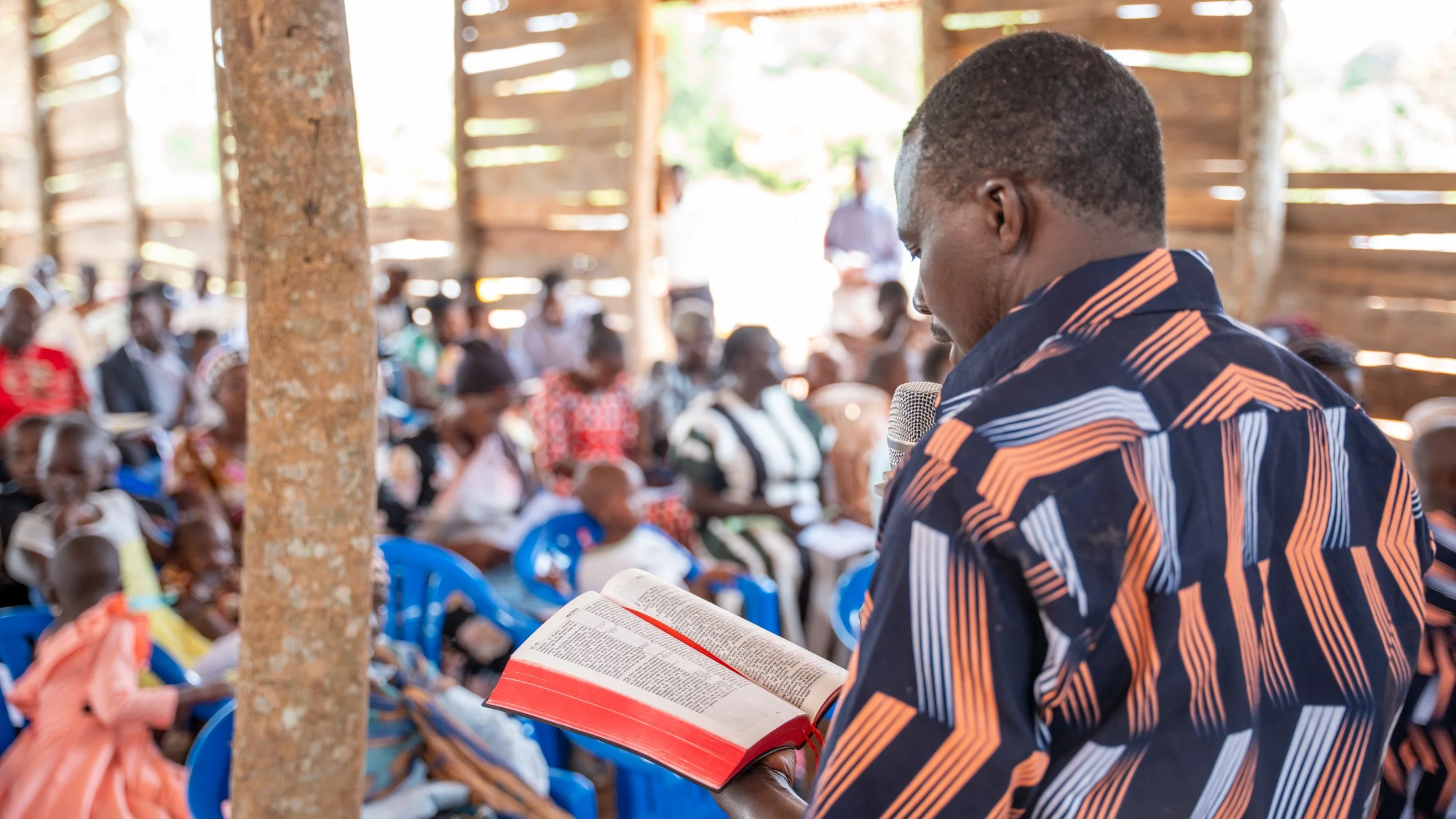 A man reading from a book during an outdoor gathering with a diverse group of people seated on blue chairs under a wooden shelter.