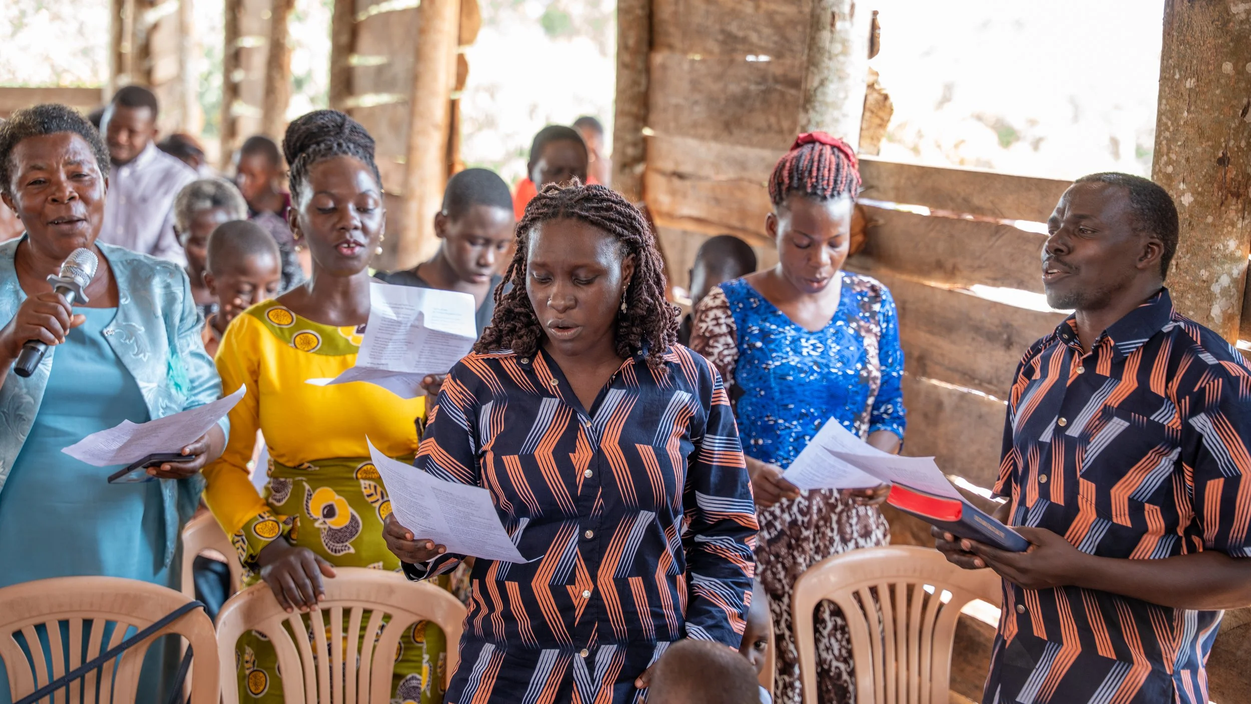 Group of people singing and reading from papers inside a rustic wooden structure.