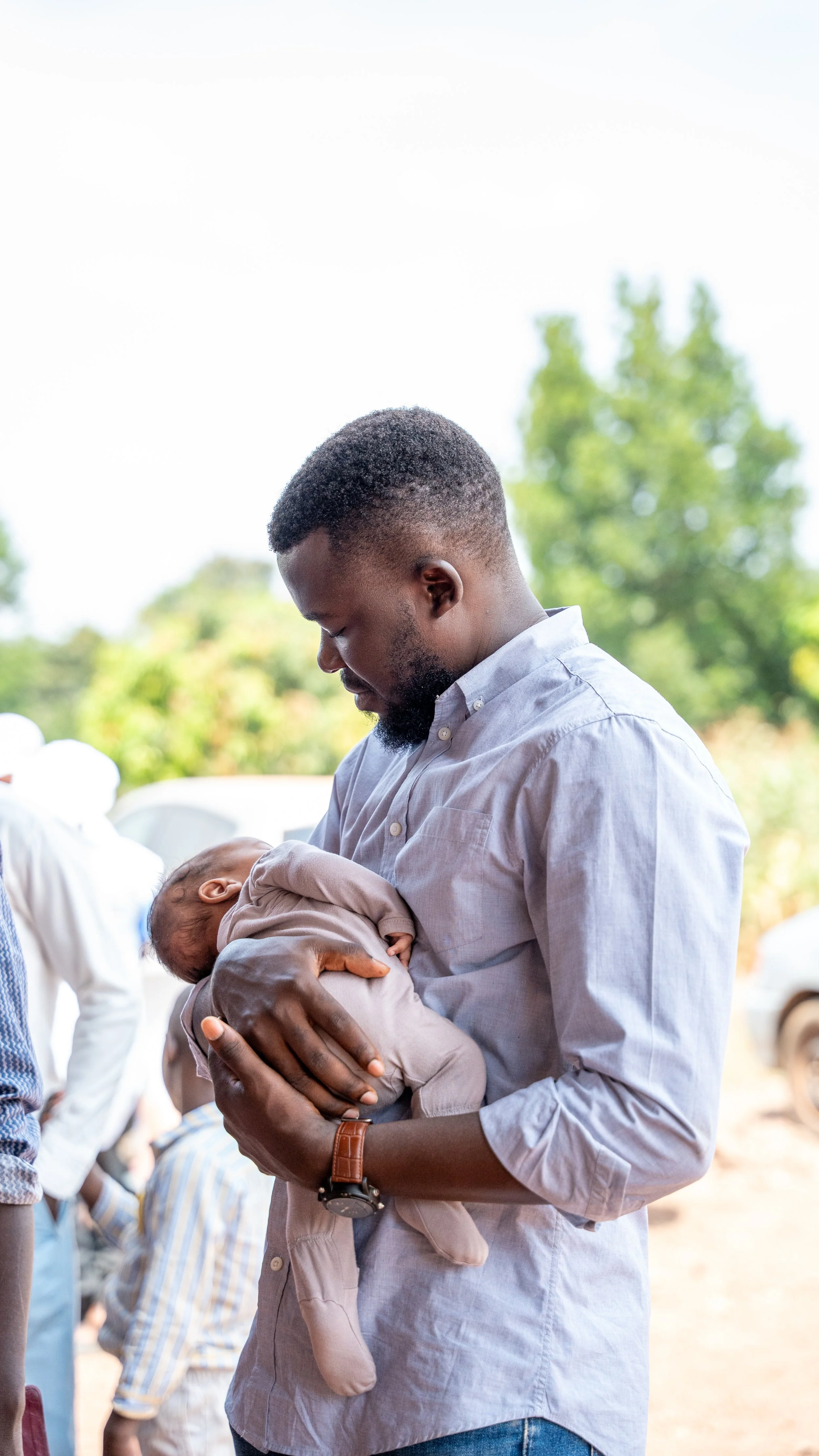 A man holding a sleeping baby outdoors, with blurred trees and other people in the background.