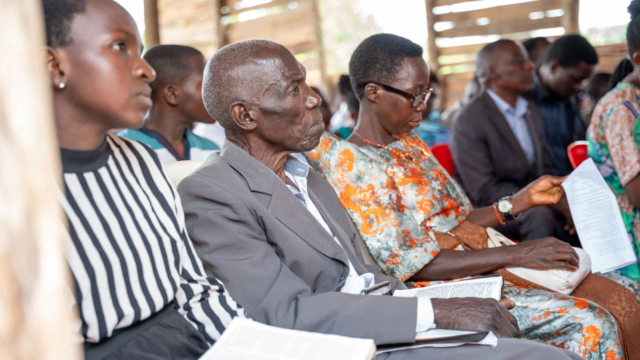 Group of diverse adults sitting in a wooden structure, attentively reading or listening during an indoor event.