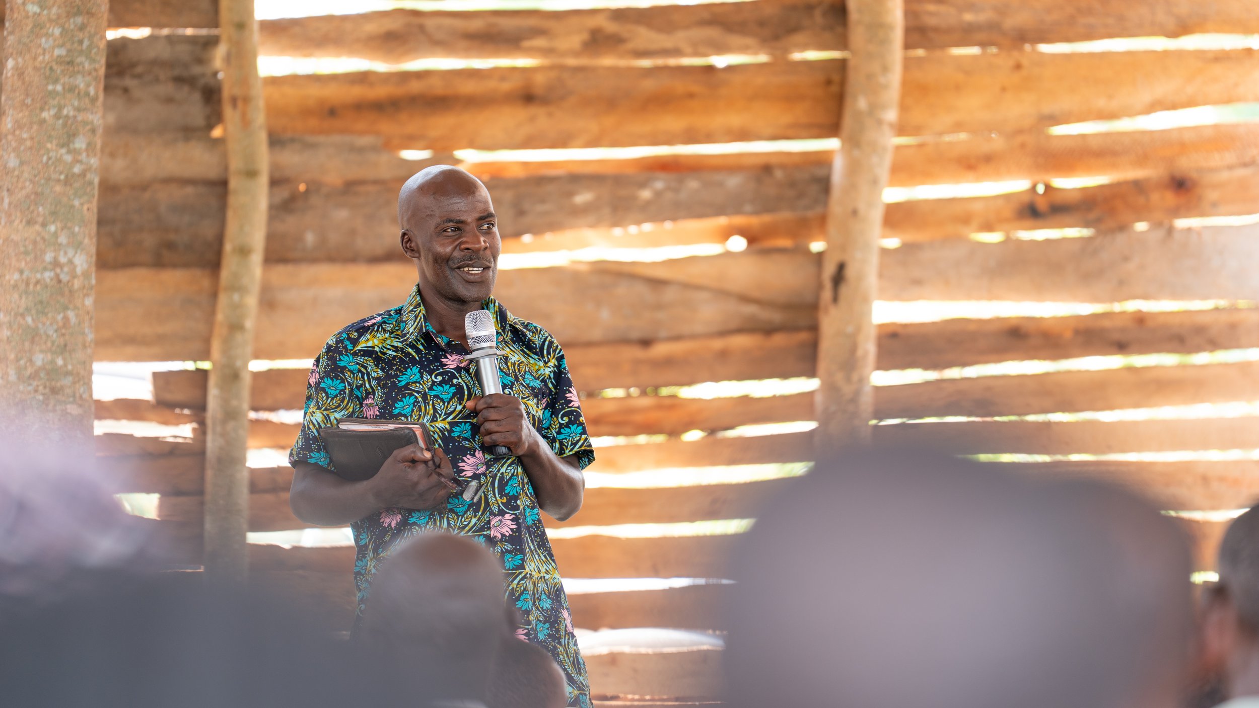 A man in a colorful tropical shirt holding a microphone and a notebook while speaking to a group indoors with a wooden wall background.