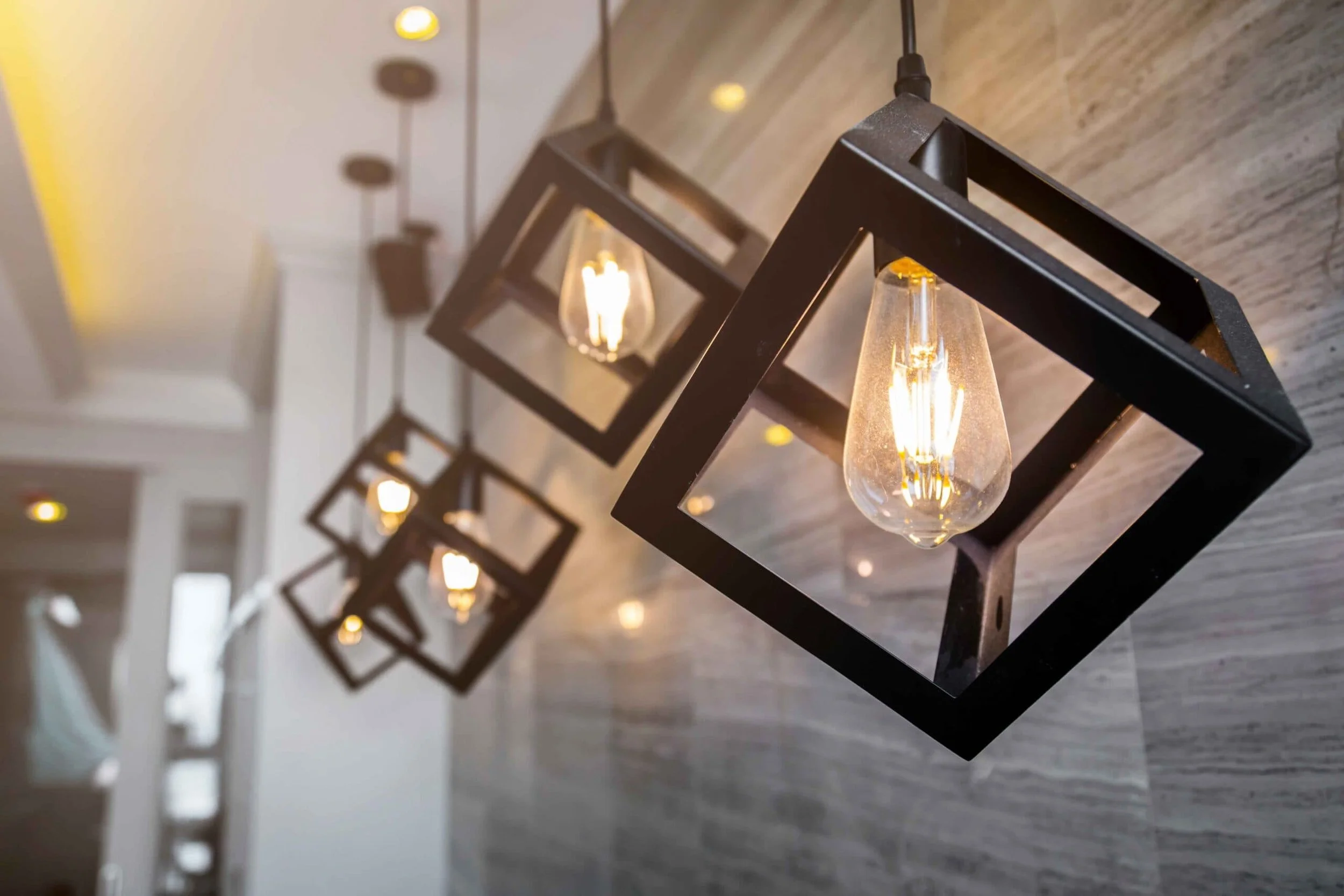 A row of modern black square-framed pendant lights with exposed filament bulbs hanging from the ceiling in a contemporary interior space.