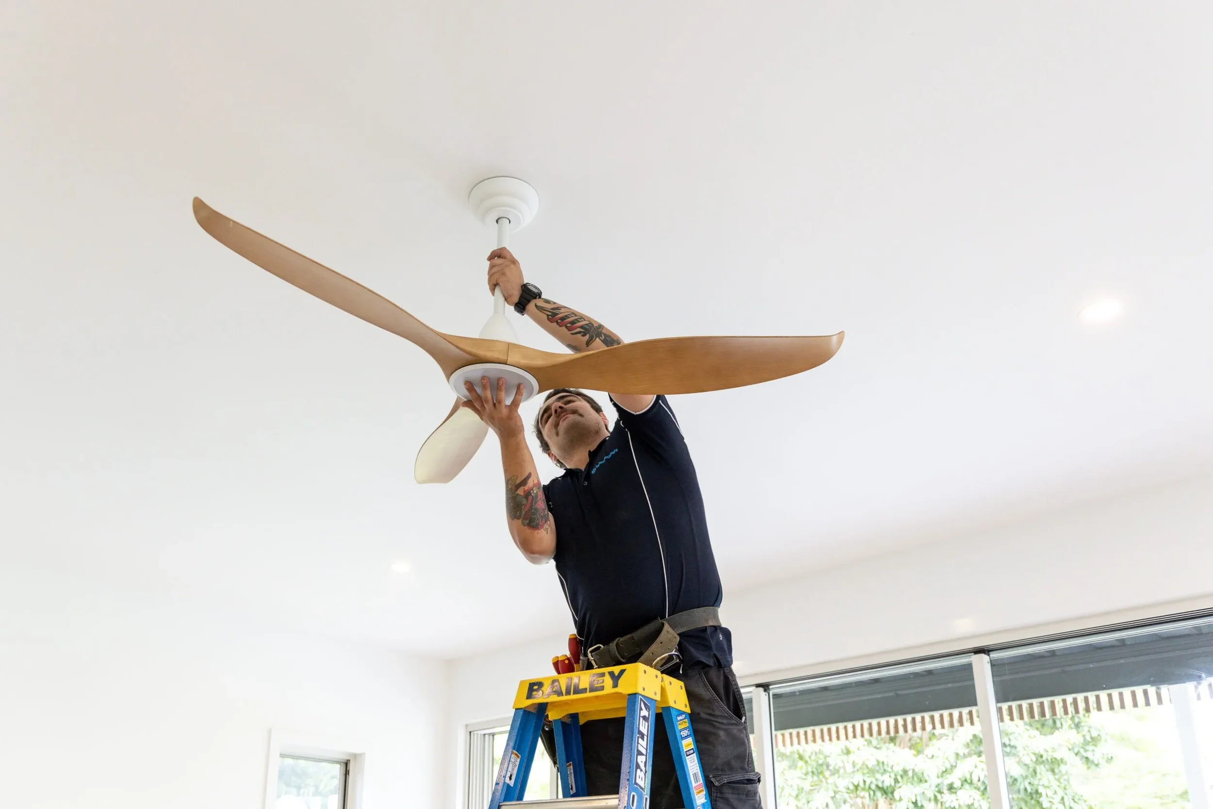 A man standing on a blue ladder installing or fixing a white ceiling fan with wooden blades in a room with large windows and white walls.
