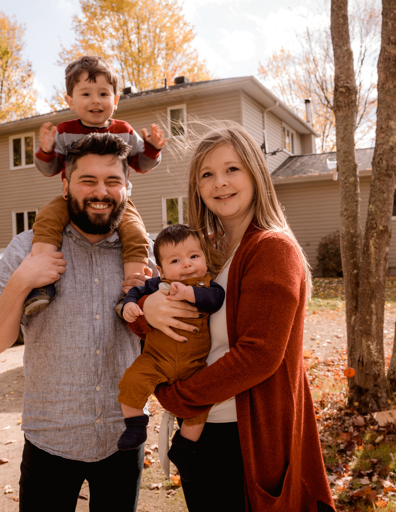 A family of four outside in autumn, with trees and a house in the background. The father is smiling and holding a toddler on his shoulders, and the mother is holding a baby. The family is dressed in fall clothing.