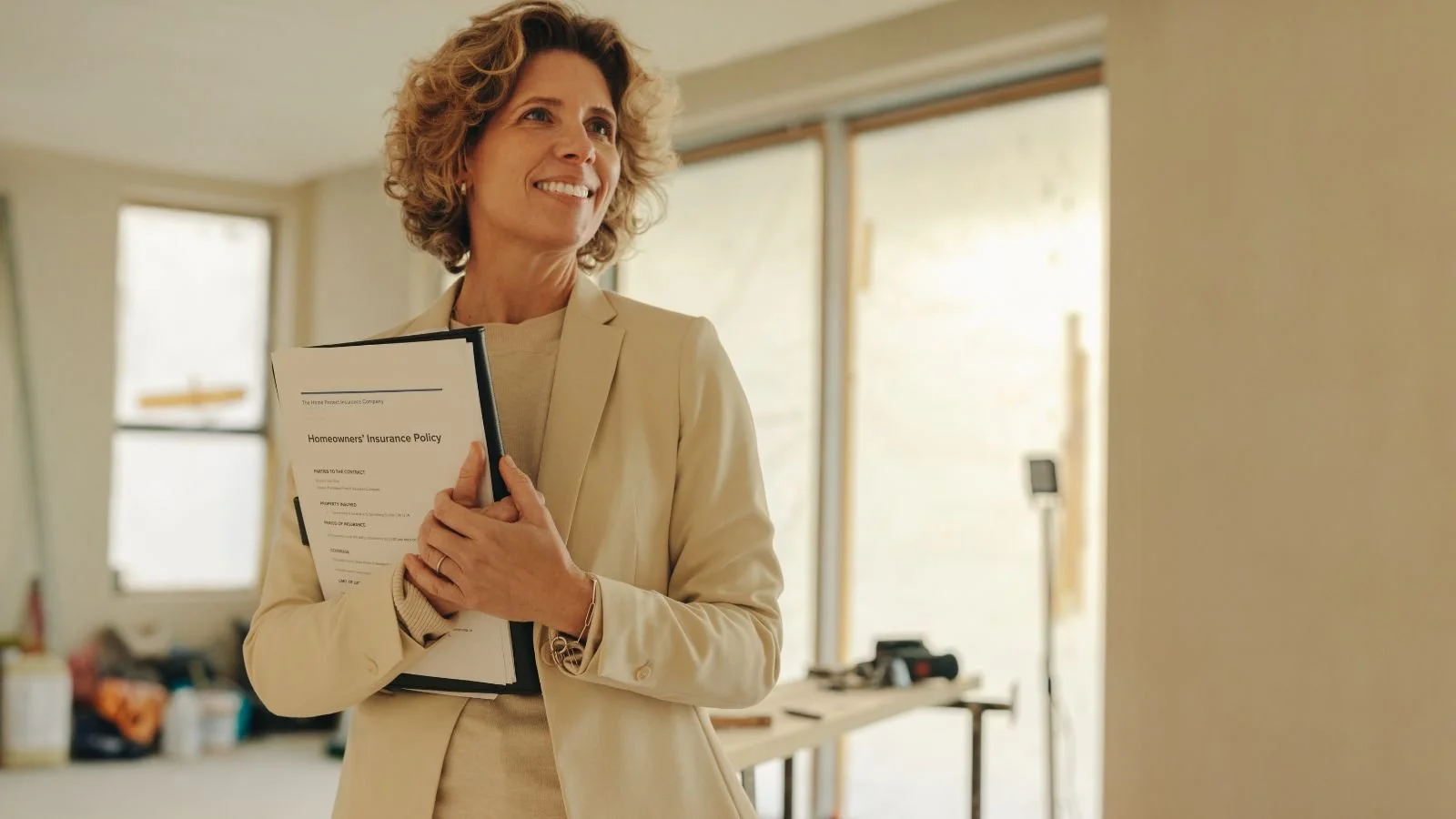 A woman with curly blonde hair in a beige blazer smiling while holding a clipboard with a document titled 'Homeowners Insurance Policy' in a brightly lit room with large windows and construction tools in the background.