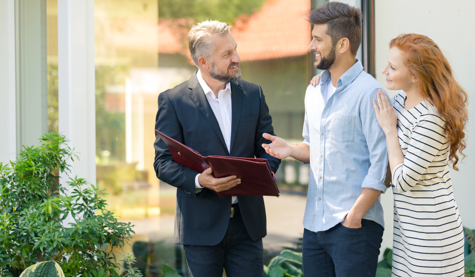 A middle-aged man in a suit holding a binder, talking to a young man and woman outside in front of a glass window, with plants nearby. The young woman has red hair and is wearing a striped dress, while the young man is wearing a light blue shirt.