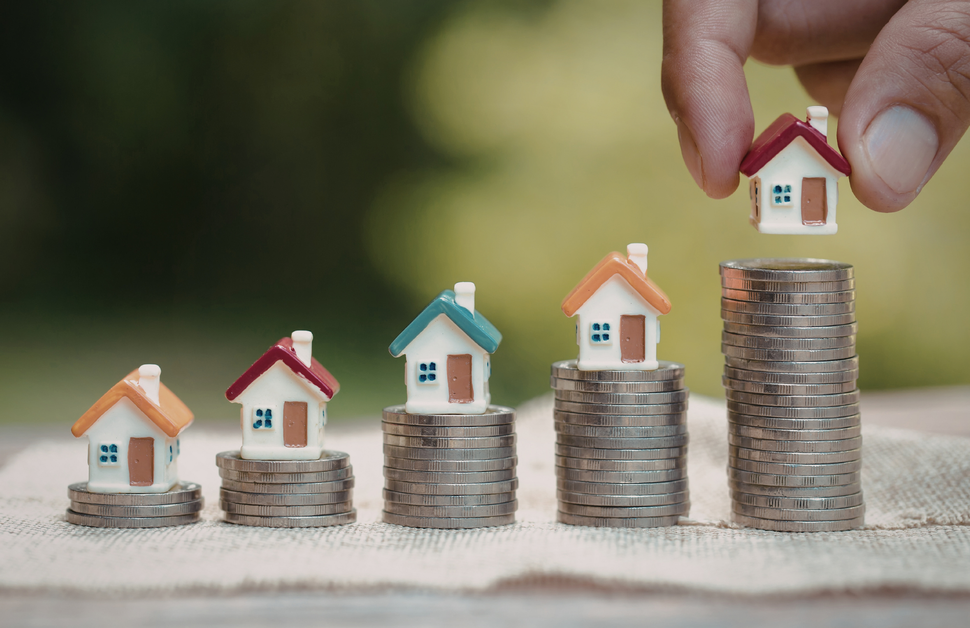 Hand placing a small house figurine on top of a stack of coins, with four stacks of coins each topped with a house figurine increasing in height from left to right.