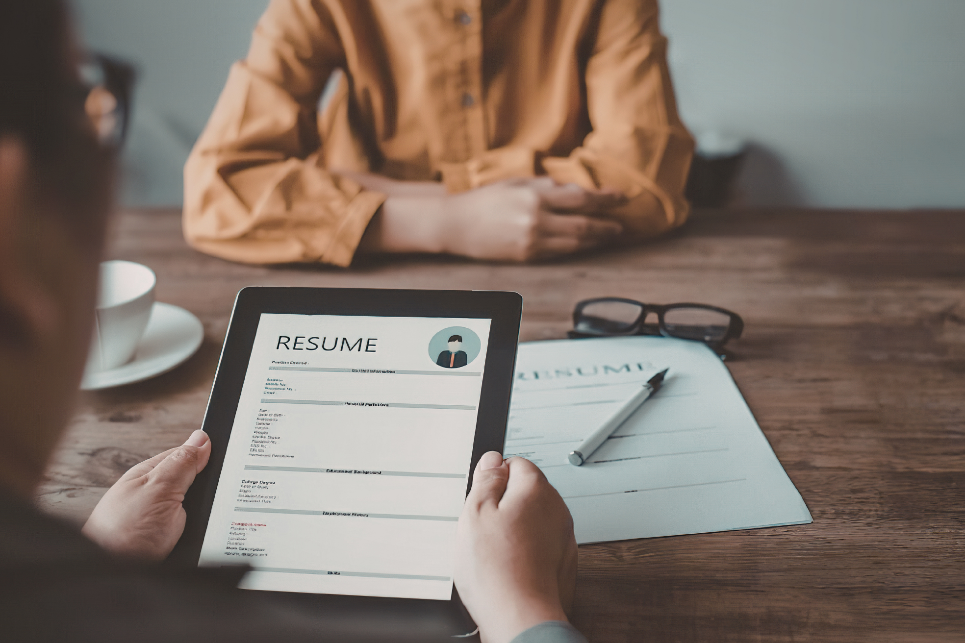 Person holding digital tablet displaying a resume, seated at a wooden table with paper, pen, glasses, and a cup of coffee.