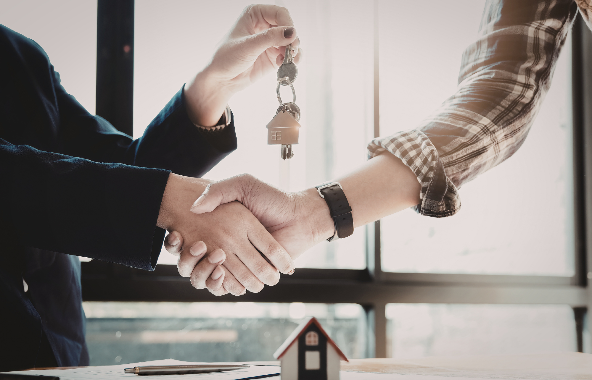 A real estate agent and a client shaking hands, with the agent handing over house keys with a house-shaped keychain.