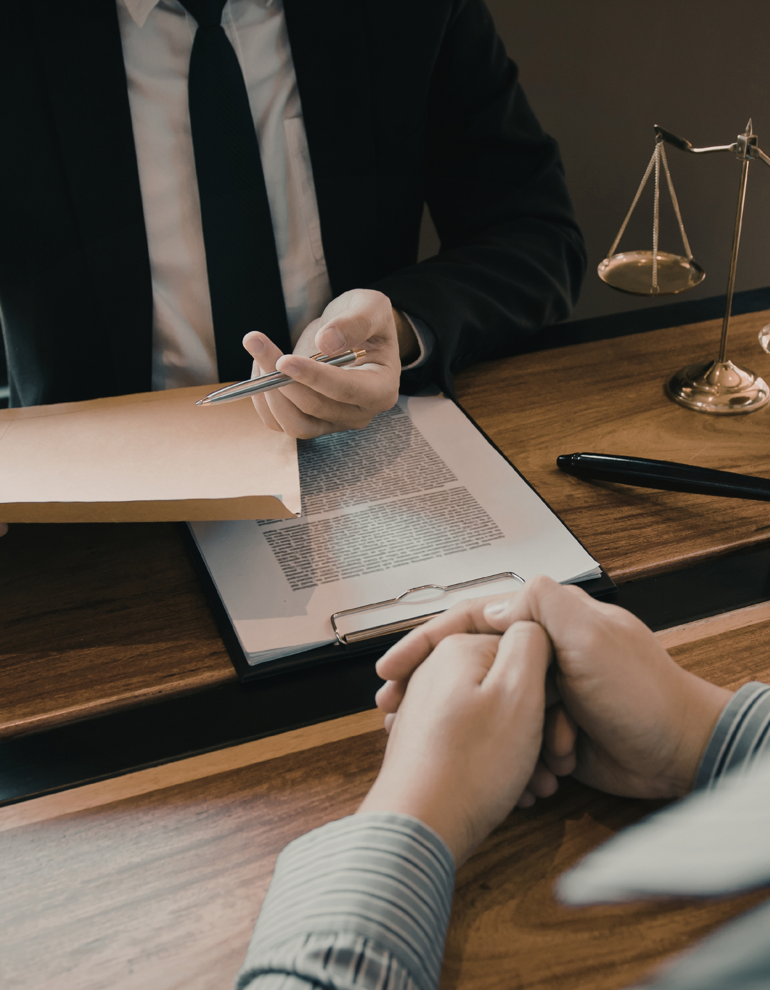 A person in a dark suit and tie sitting at a wooden desk, holding a pen over documents. Another person with clasped hands sits across the desk. A small scales of justice statue is on the desk.