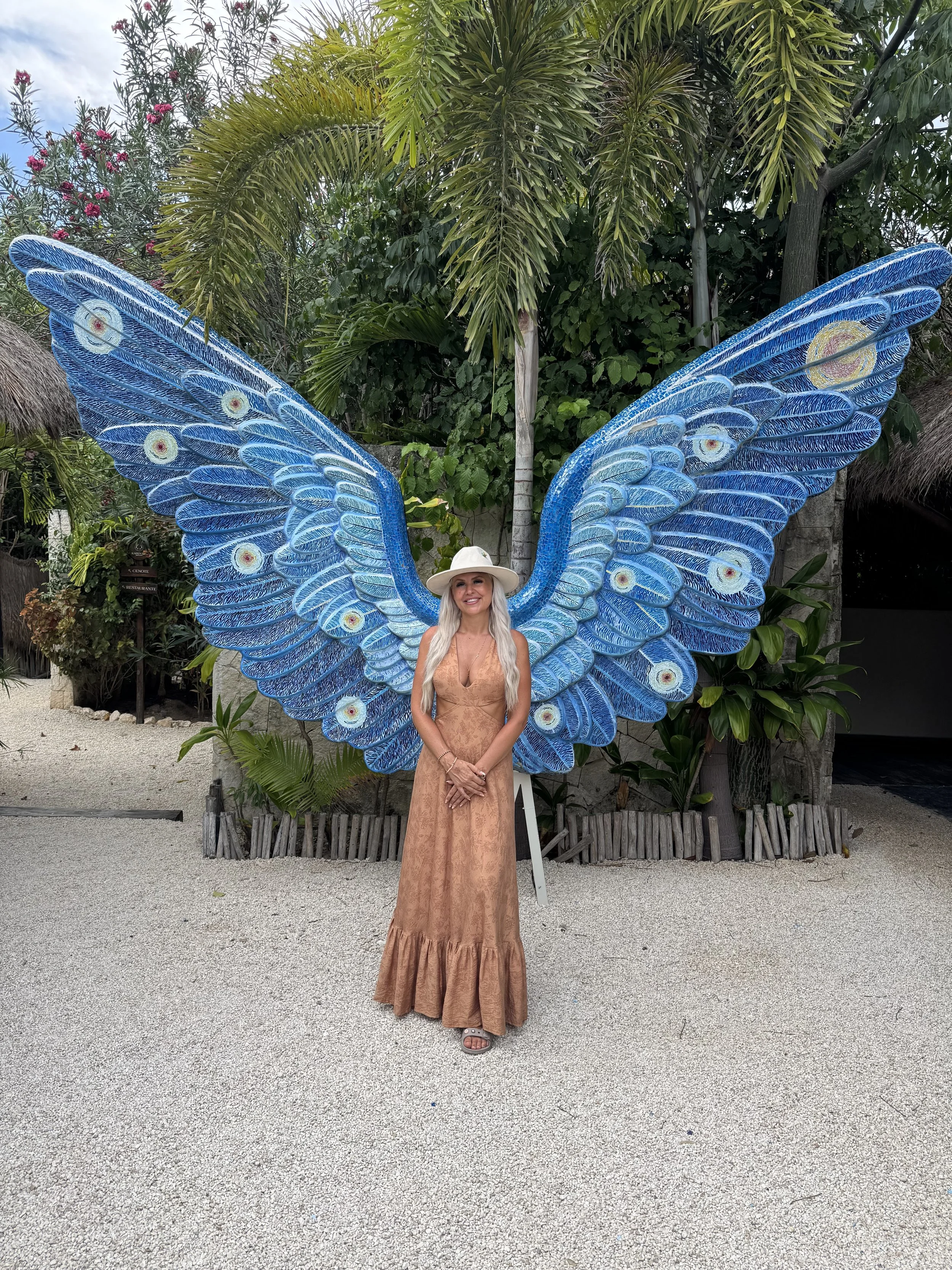A woman standing outdoors in front of a large butterfly decoration, with green tropical plants and a thatched-roof structure in the background. She is wearing a long, tan-colored dress and a white wide-brimmed hat.