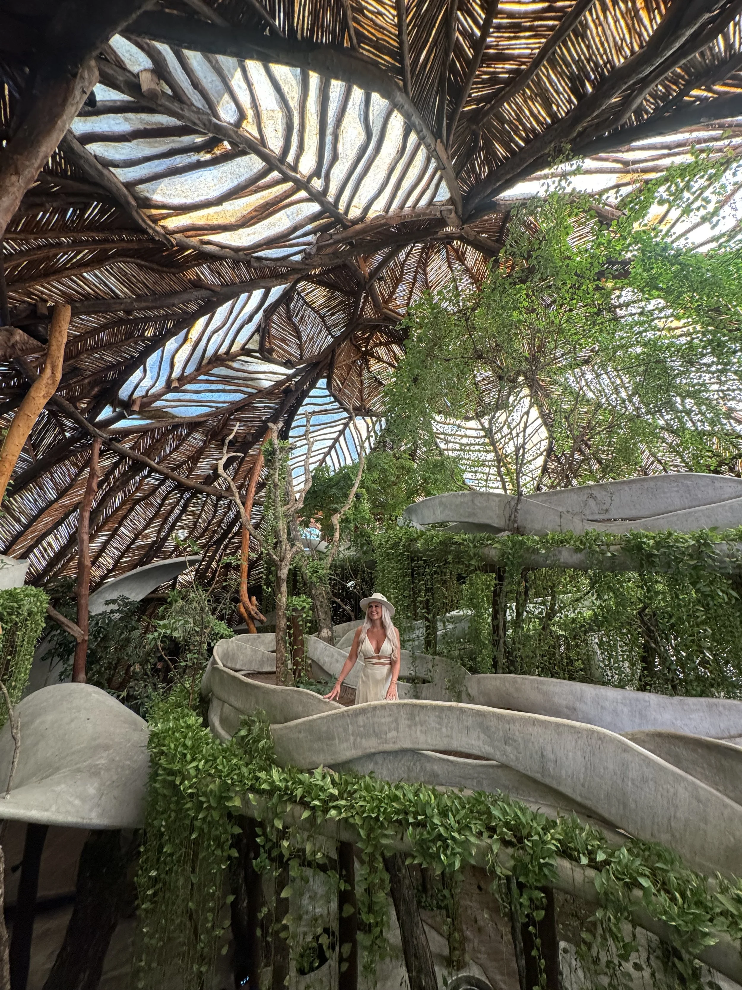 A woman in a white dress and hat is standing among lush green plants inside a structure with wooden and thatched roofing, surrounded by curved concrete planters.