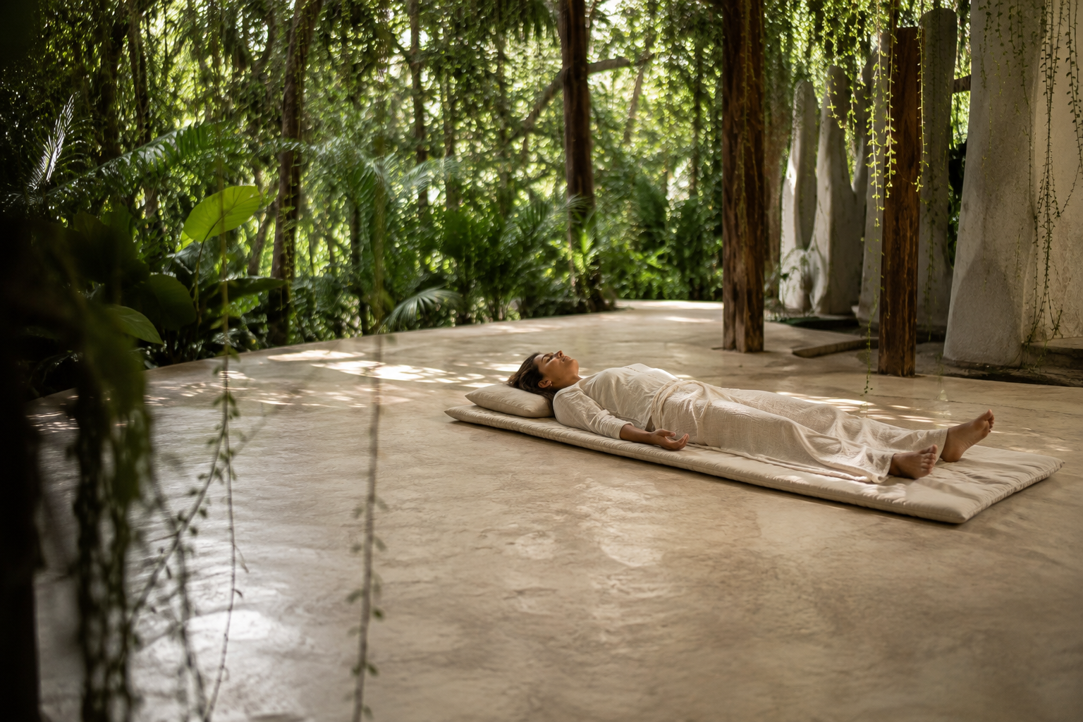 A woman in white loungewear lying on a mat on the floors of a lush, green, open-air space with hanging plants and wooden pillars.