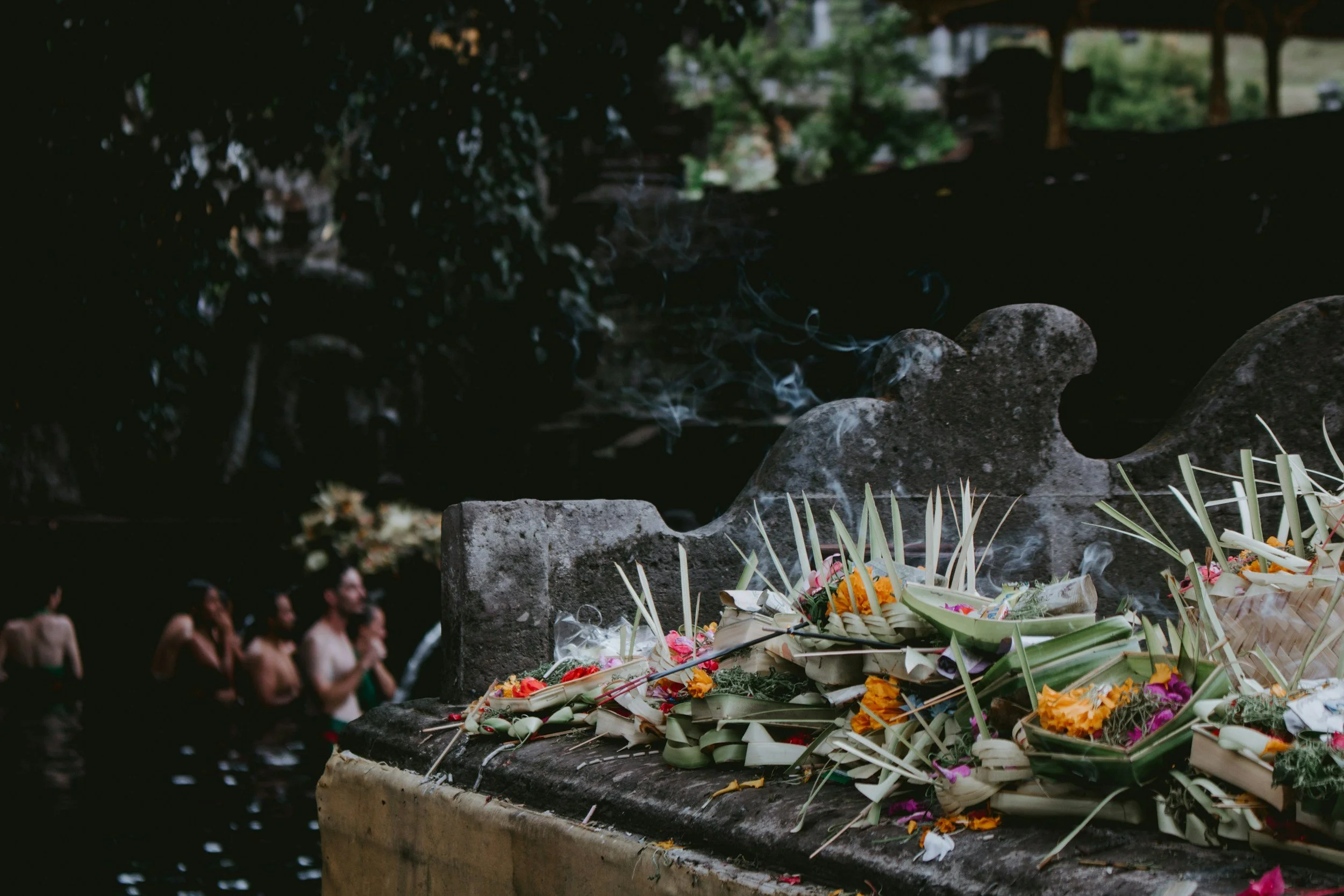 Relief am Flussufer mit Blumen und Räucherstäbchen, Menschen beim Ritual in einem Tempel im Hintergrund.