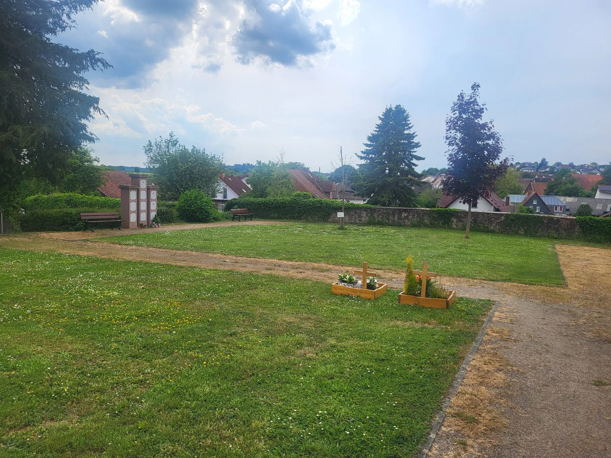 Ein Park mit Bänken, Blumenbeeten und Bäumen, im Hintergrund ein Dorf mit Häusern und Bäumen. Der Himmel ist bewölkt.