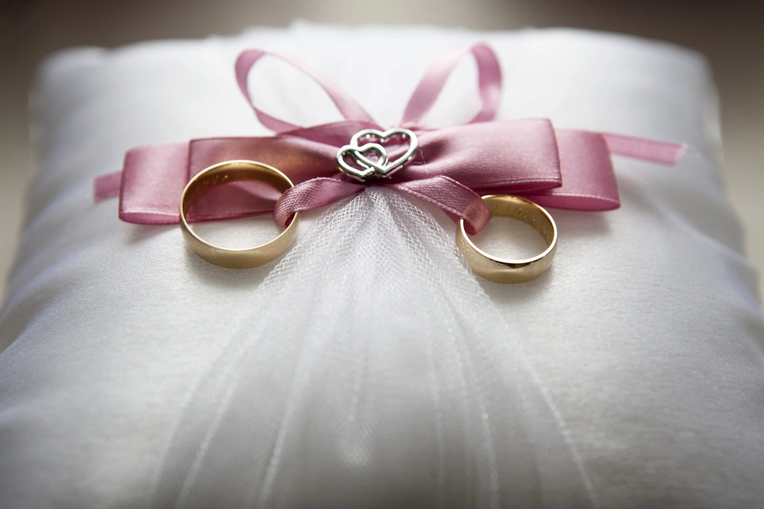 Ein Geschenk mit weißen Stoff, verziert mit rosa Schleifen, zwei goldene Eheringe und ein silbernes Herz-Schloss als Dekoration.