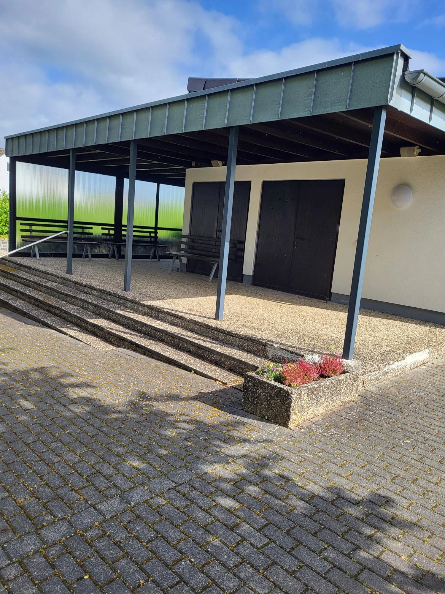 Ein moderner, überdachter Eingangsbereich mit Sitzbänken, Treppen und Blumenbeet vor einem Gebäude. Das Gebäude hat schwarze Türen und eine weiße Wand.