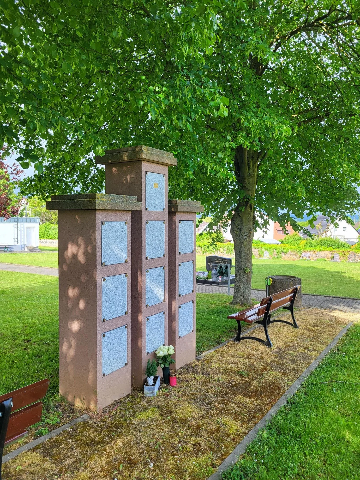 Ein Grabmal mit mehreren Tafeln in einem Park unter einem Baum, mit Blumen und Kerzen davor.