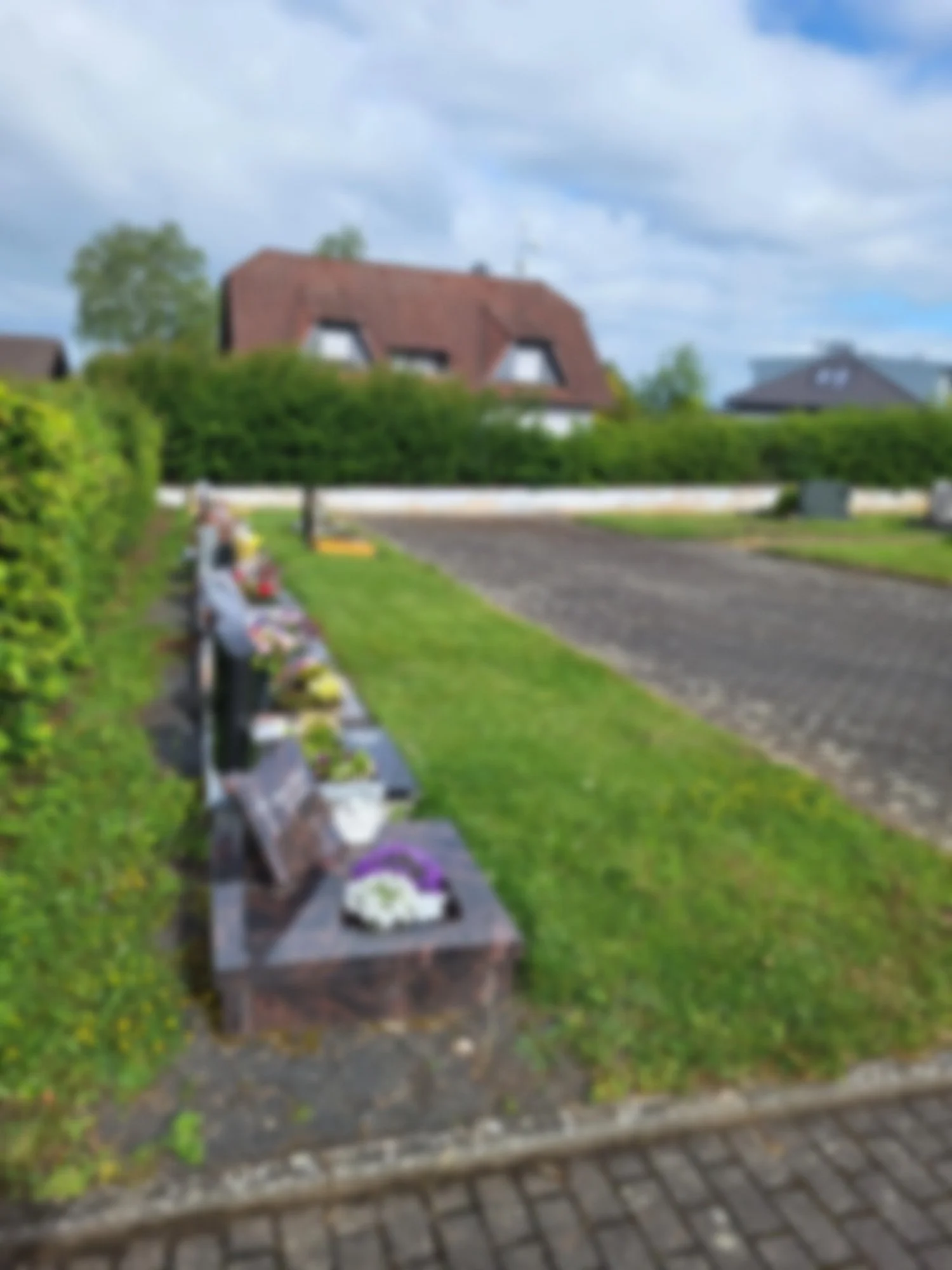Blurry Foto eines Friedhofs mit Grabsteinen und einem Blumenarrangement, im Hintergrund Häuser und Bäume bei bewölktem Himmel.