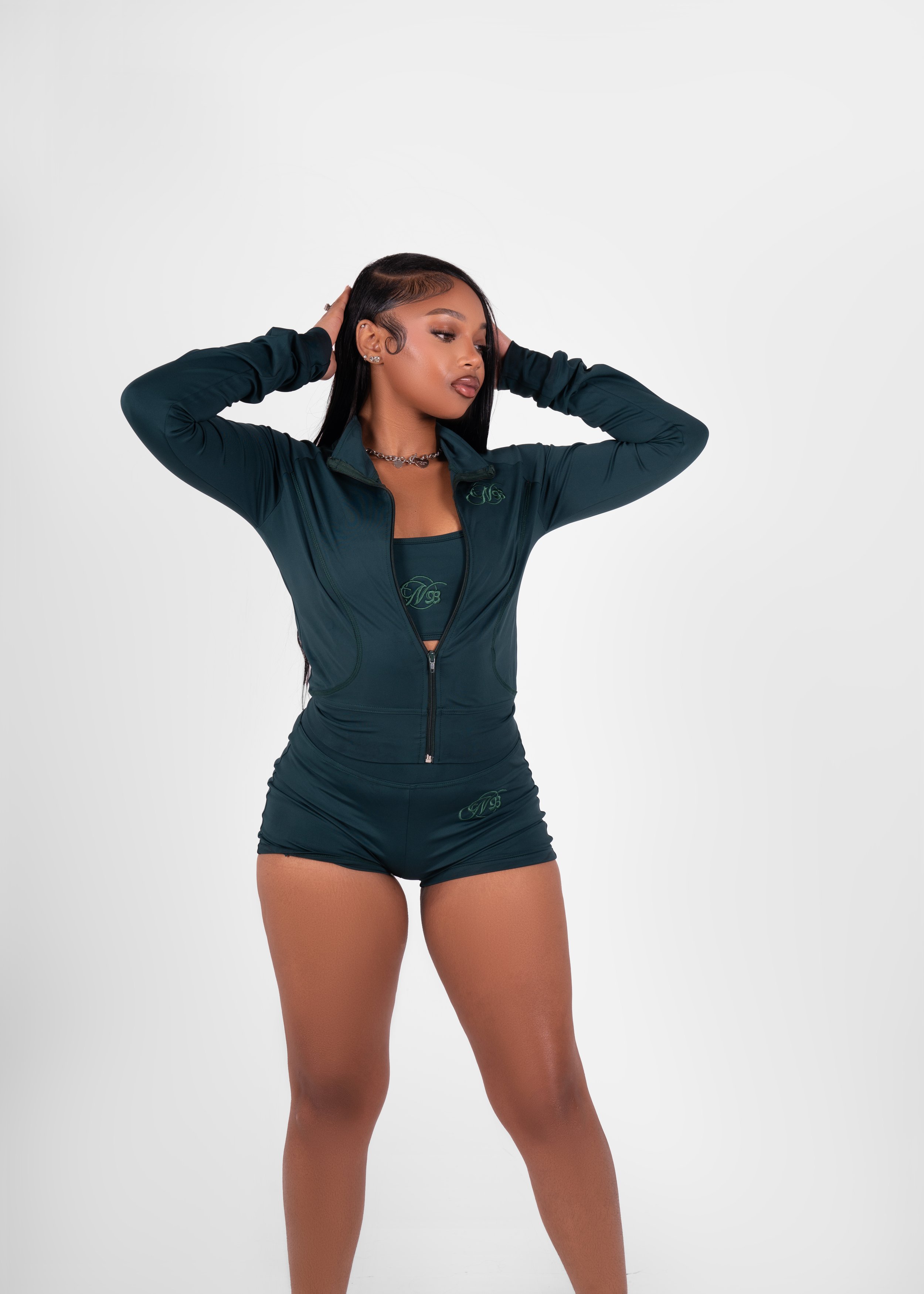 A woman wearing a dark teal athletic jacket and matching shorts, standing with her hands behind her head, against a plain white background.