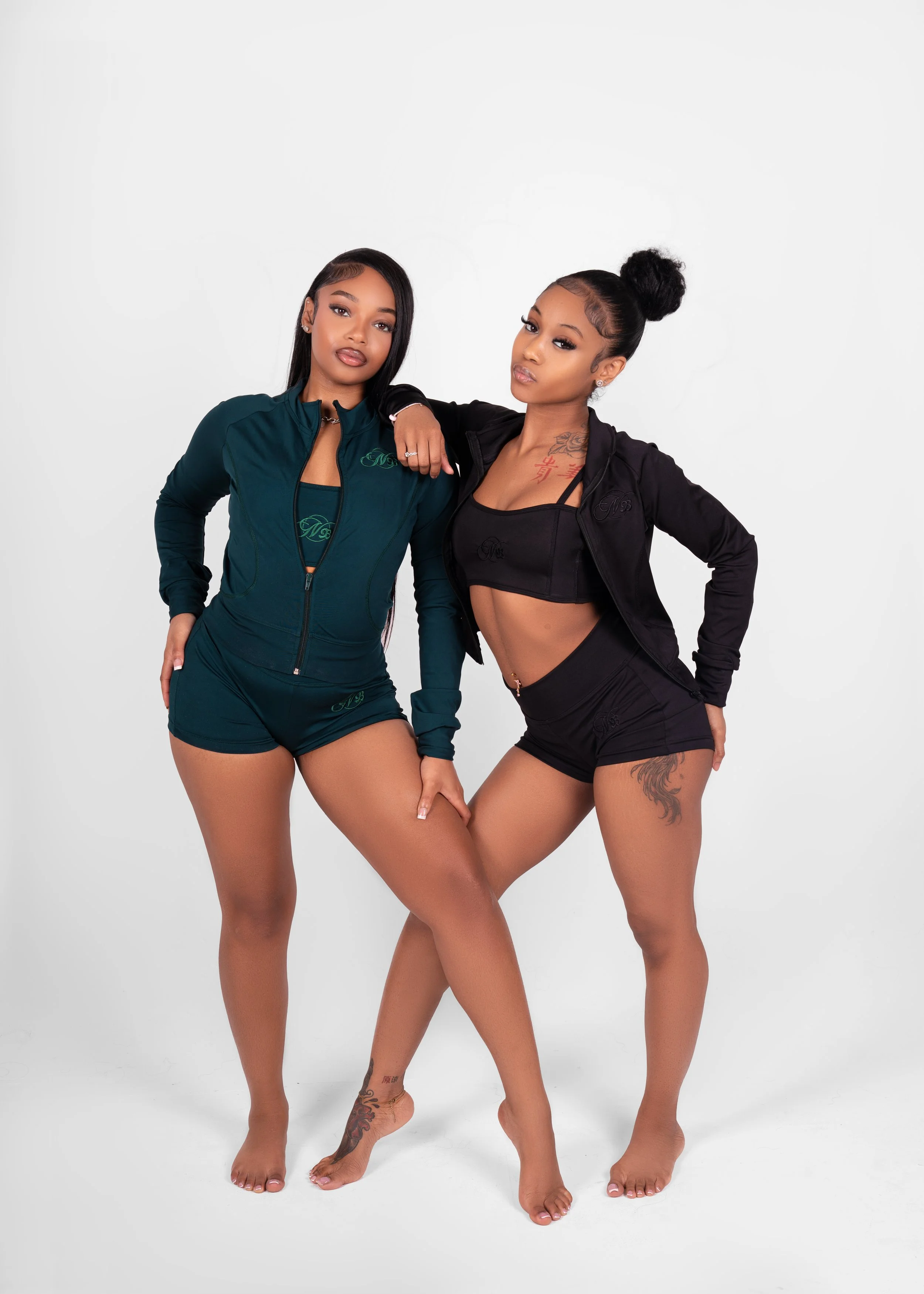 Two women standing barefoot against a white background, wearing athletic outfits, with one woman resting her arm on the other's shoulder.