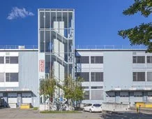 Ein modernes Gebäude mit einer Glaseinhaussbrücke und mehreren Stockwerken, umgeben von Bäumen und einem Parkplatz.