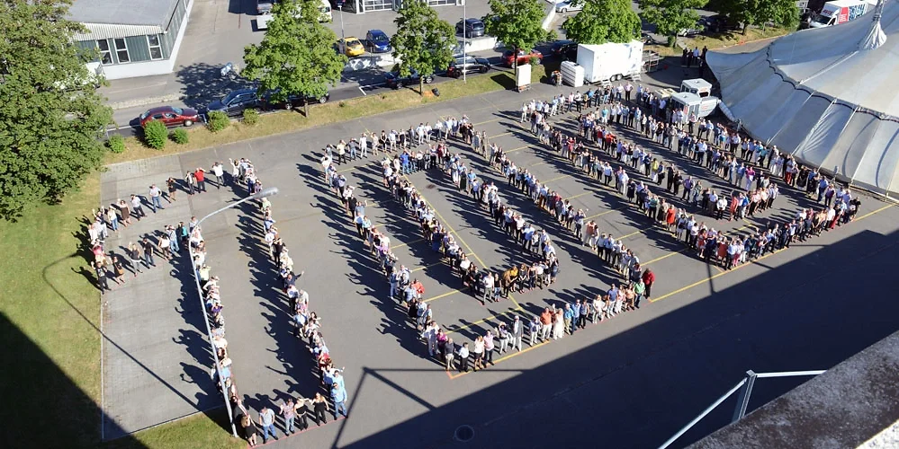 Menschen stehen in einer langen Schlange auf einem Schulhof, die die Worte 'COVID' in Großbuchstaben bildet, umgeben von Bäumen und Gebäuden, bei gutem Wetter.