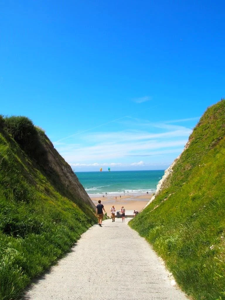 A paved walking path cutting through lush green hills leading down to a wide sandy beach and turquoise sea on the Opal Coast in France.