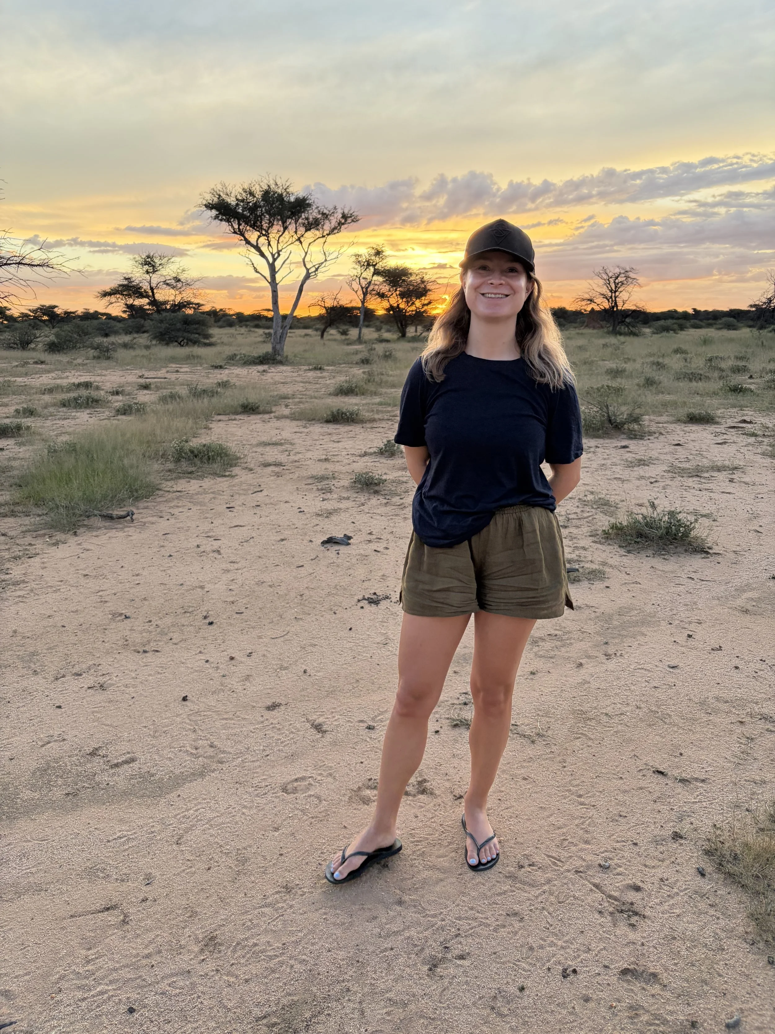 Smiling woman in black t-shirt, shorts, and flip-flops stands in a desert landscape at sunset with sparse trees and a colorful sky.
