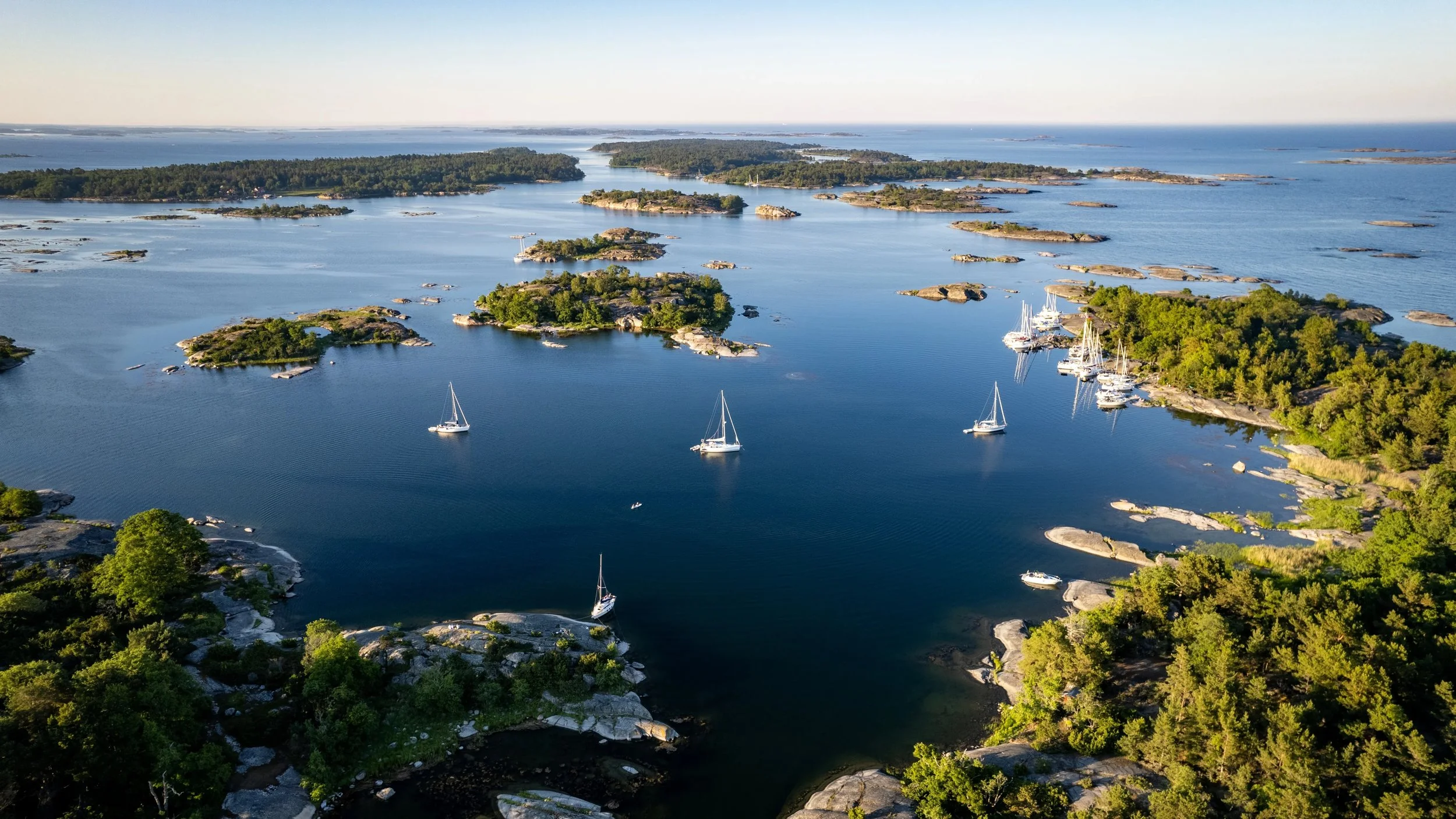 Aerial drone view of sailboats cruising through the small, rocky green islands of the Stockholm Archipelago in Sweden under a clear blue sky.