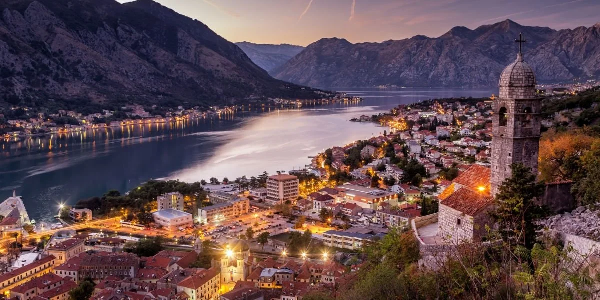 A stunning twilight view of Kotor, Montenegro, with the old town lights glowing along the winding bay, looking out from a stone church tower toward the dark mountains.