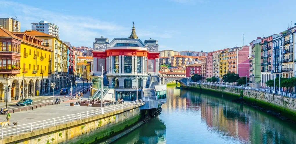 The scenic Nervión River waterfront in Bilbao, showing the historic Ribera Market alongside vibrant, colorful buildings reflecting in the water.