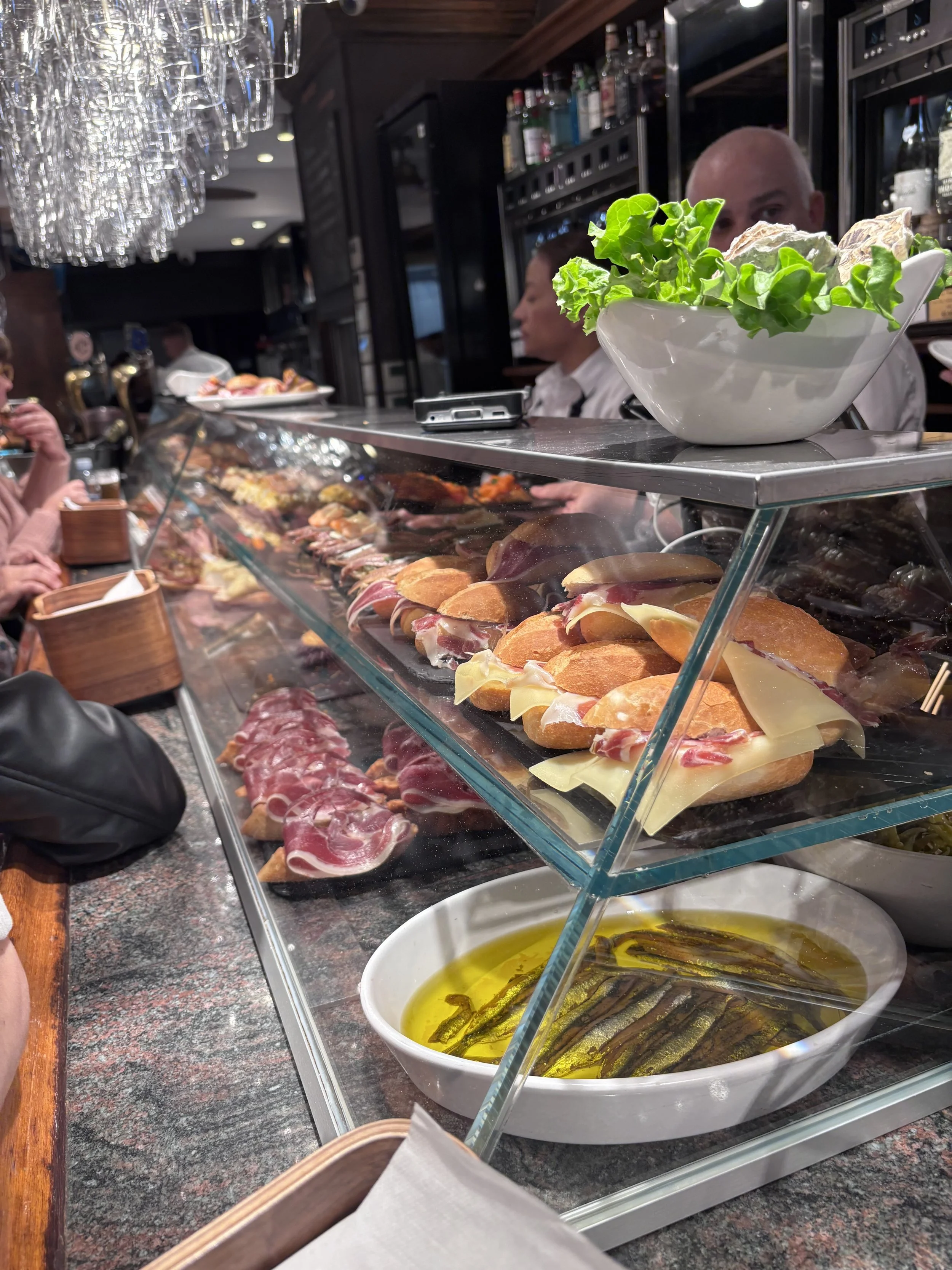 A glass counter display inside a traditional Basque bar packed with authentic pintxos, including cured Iberico ham and cheese baguette sandwiches.