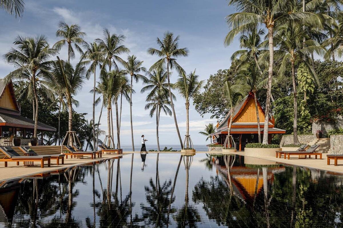 A luxury infinity swimming pool at a tropical resort in Thailand, surrounded by tall palm trees reflecting in the still water with ocean views.