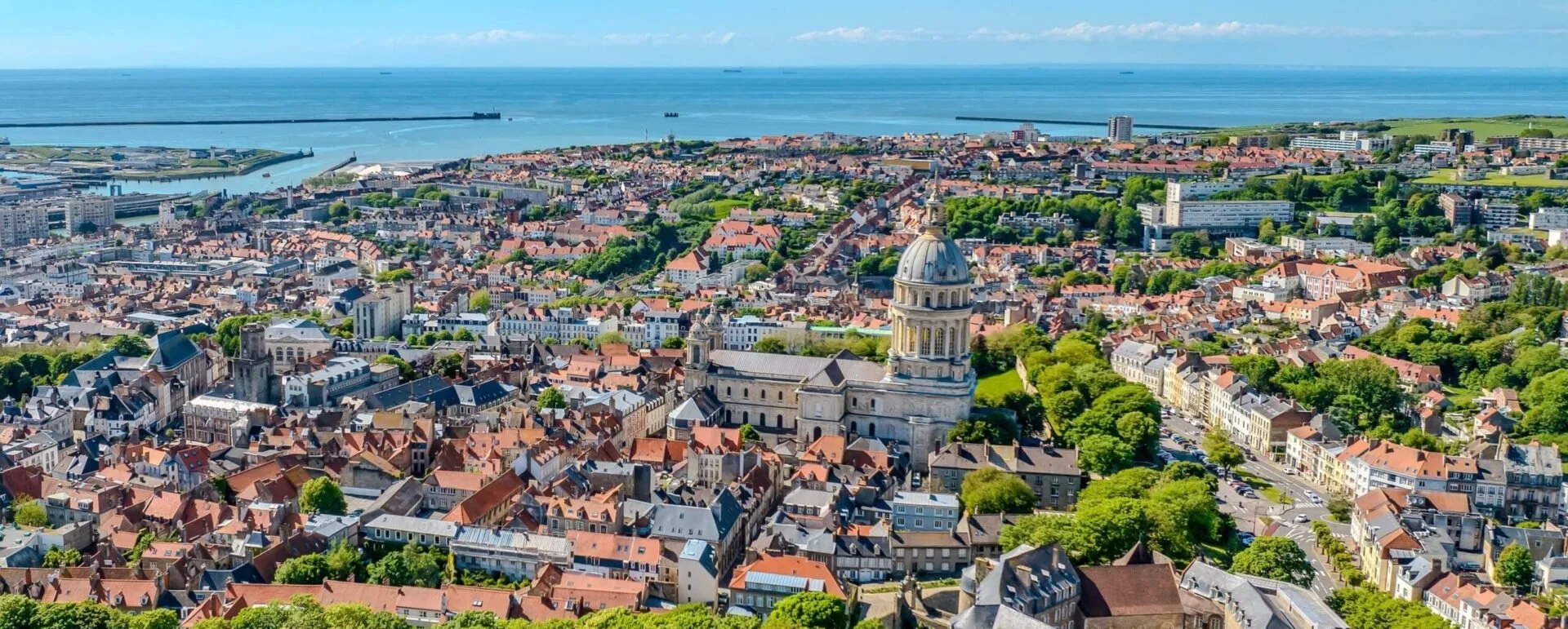 An aerial panoramic view over the historic red-roofed buildings and grand cathedral dome of Boulogne-sur-Mer next to the blue ocean.