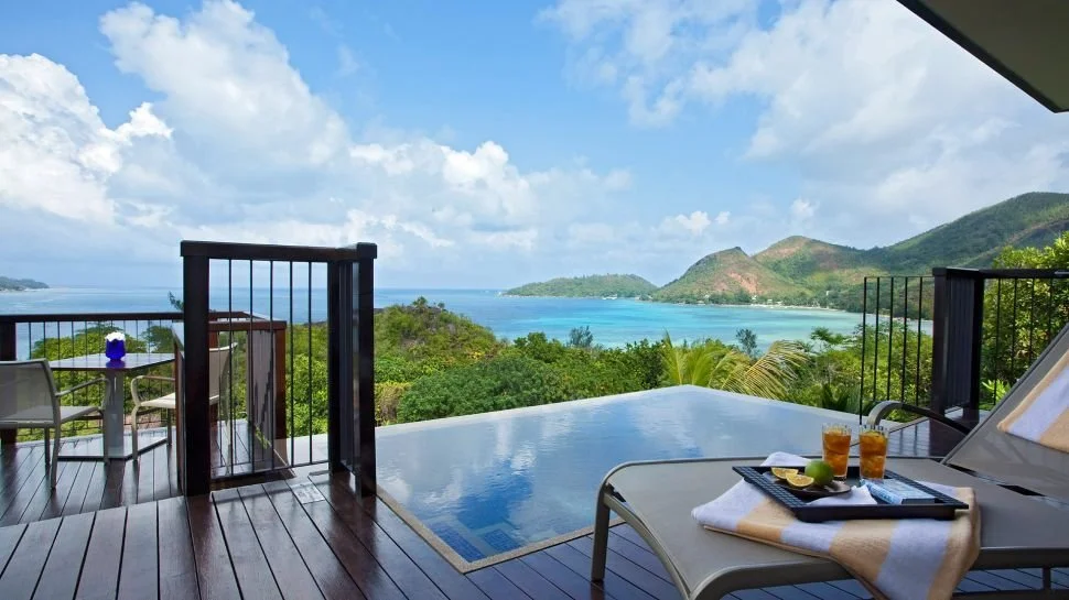 A private luxury villa balcony in the Seychelles featuring an infinity plunge pool, sun loungers with drinks, and a panoramic view of the turquoise Indian Ocean.