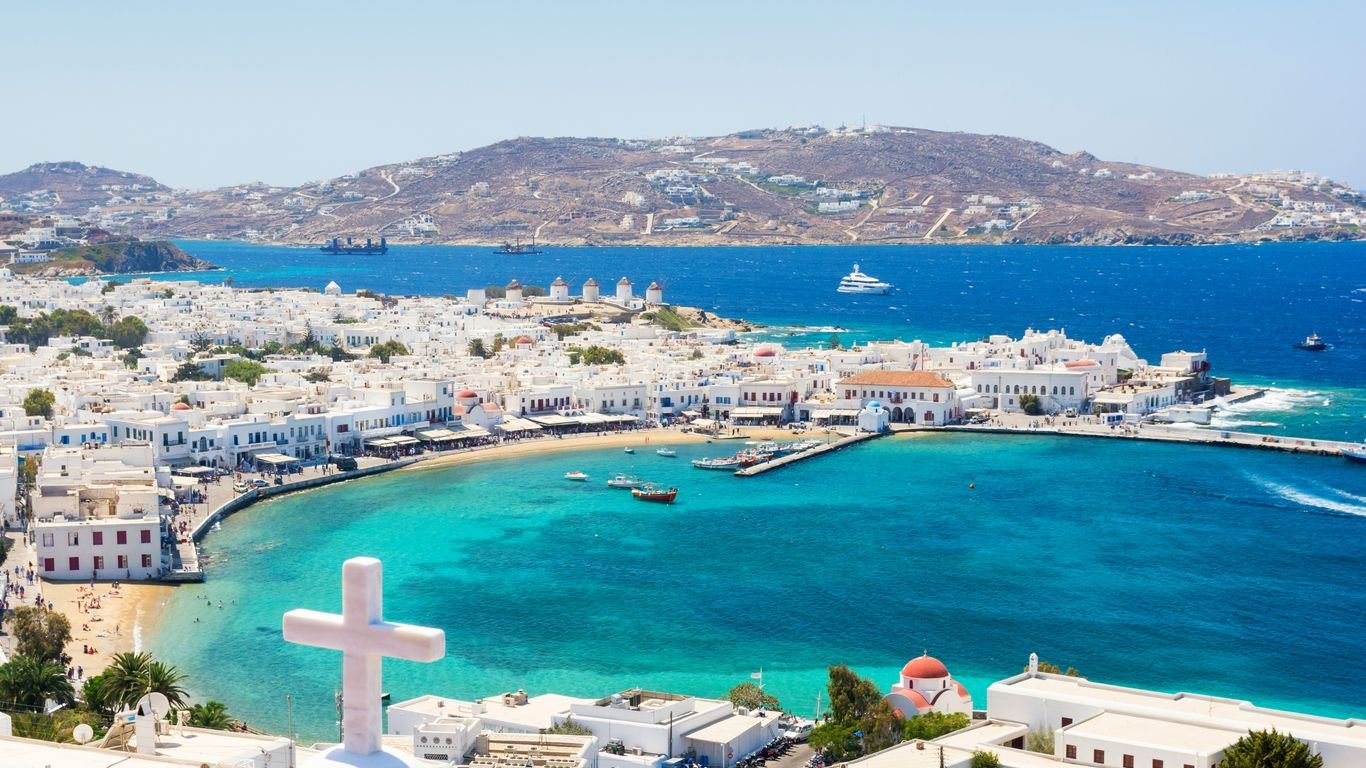 High-angle panoramic view of a coastal Greek island town featuring iconic white buildings, a turquoise bay with boats, and a large white cross in the foreground.