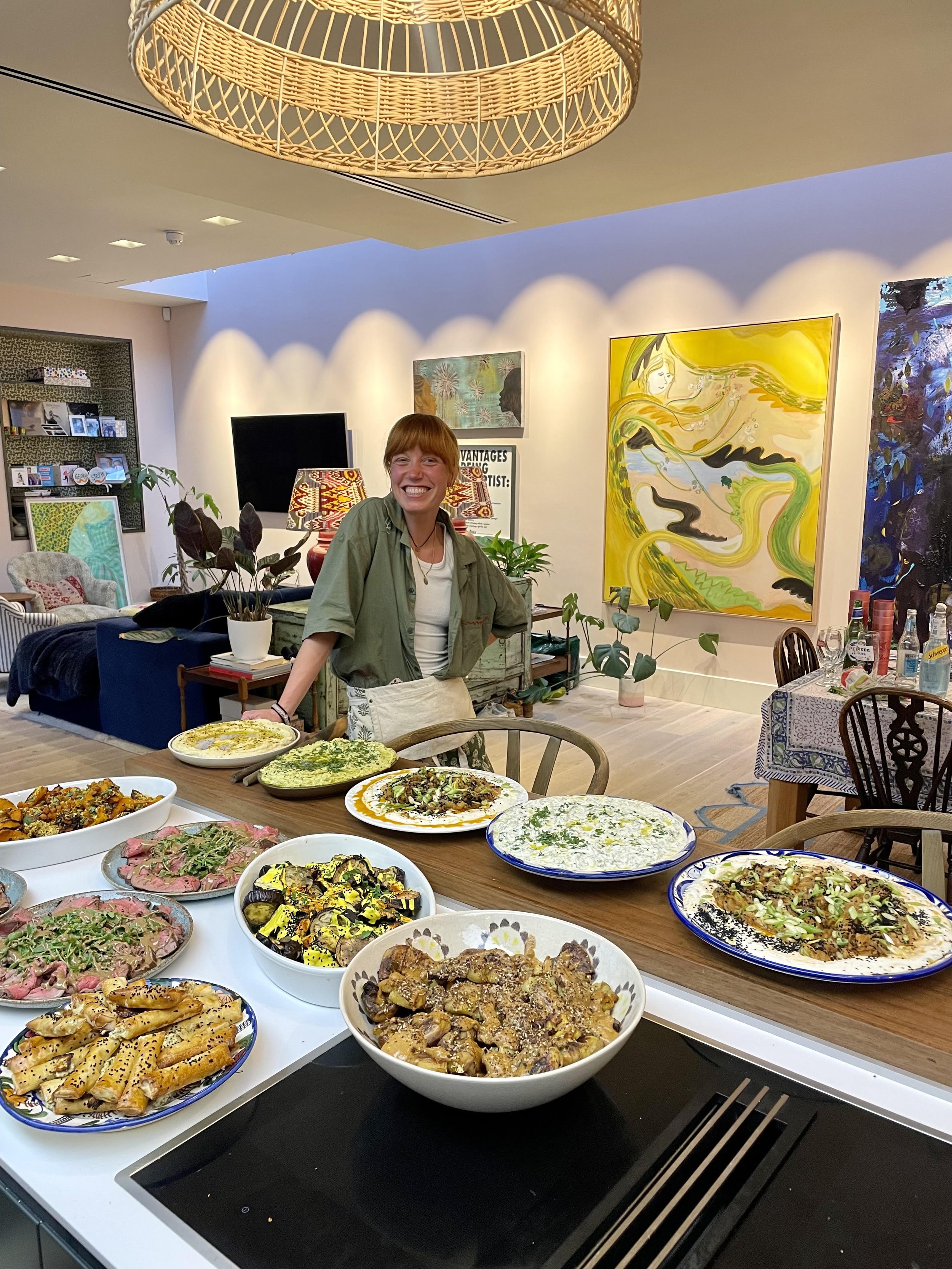 A woman with short red hair and a green shirt standing behind a kitchen counter with various dishes, smiling. The background features a modern, art-filled living space with plants, a television, and colorful abstract paintings.