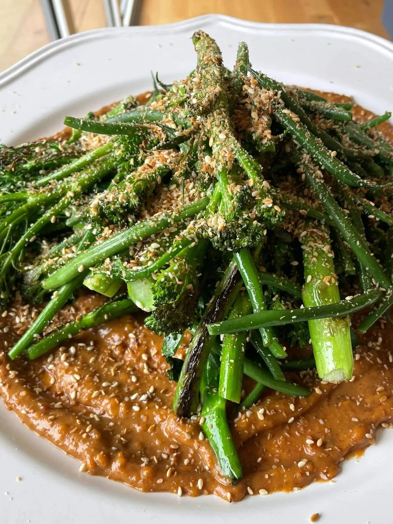 A plate of cooked green vegetables, such as broccolini and green beans, topped with sesame seeds and crushed nuts, served over a smear of brown sauce.
