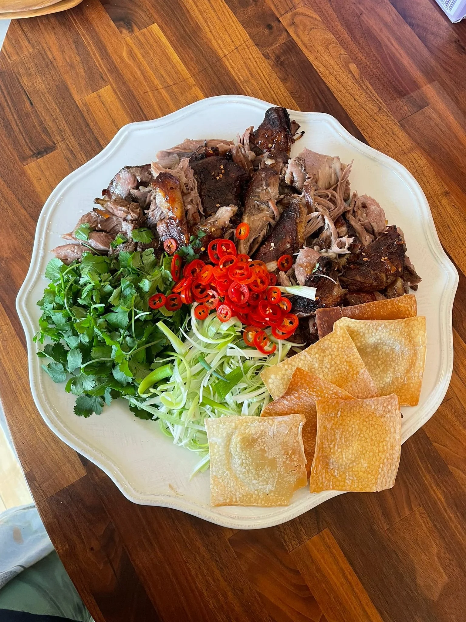A large white plate with roasted meat, fresh cilantro, sliced red chili peppers, shredded green onions, and fried tofu skins on a wooden table.