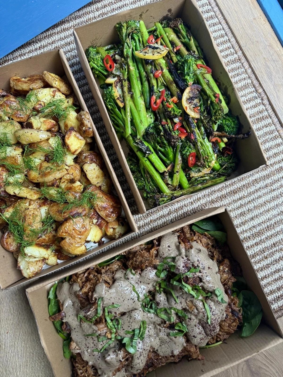 Three separate boxes of food on a woven placemat. The top box contains grilled asparagus with lemon slices and red chili peppers. The left box has roasted fingerling potatoes garnished with herbs. The bottom box holds a meat dish with gravy, topped with chopped green onions.